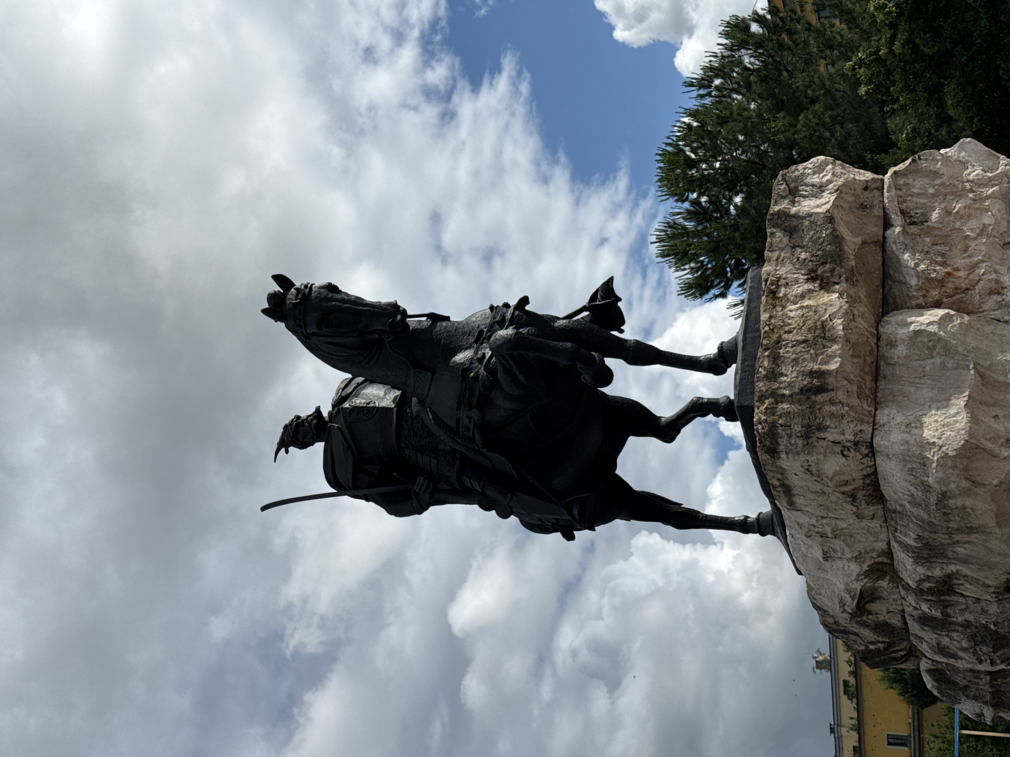 The image displays an equestrian statue, captured from a low angle, set against a bright, partly cloudy sky. The central subject is a statue of a historical figure, likely a warrior, mounted on a horse. Both the rider and the horse are rendered in a dark, possibly bronze or iron, material, appearing as a silhouette against the sky. The rider is depicted holding a sword, while the horse is in a dynamic, rearing pose, with its front legs lifted.
The statue is elevated on a substantial pedestal made of large, rough-cut stone blocks, which are light in color. In the background, to the right of the monument, dense green trees are visible. Beyond the trees, glimpses of a light yellow building with dark windows can be seen. The sky shows patches of clear blue interspersed with large, white, fluffy clouds, indicating a bright day.
The scene suggests an outdoor public space, such as a square or park. Given the location context, this is the Skanderbeg Monument in Tirana, Albania, commemorating the national hero Gjergj Kastrioti Skanderbeg. No visible text is present on the statue or its base. The image conveys a sense of historical significance and grandeur.