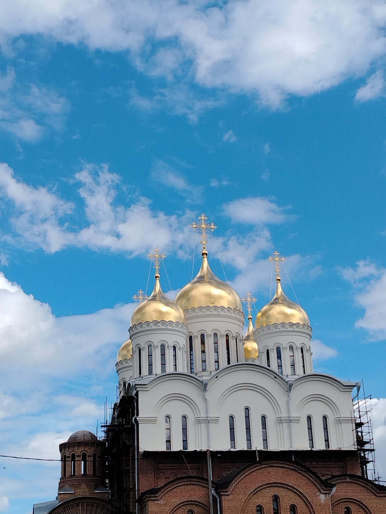 The image depicts the Assumption Cathedral (Uspensky Sobor) in Yaroslavl, Russia. The cathedral is characterized by its multiple golden domes topped with crosses, contrasting against a bright blue sky dotted with white clouds. The lower portion of the building is constructed of red brick, with white architectural details and numerous arched windows. Scaffolding is visible on the right side of the building, suggesting ongoing construction or renovation. The scene is focused on the architectural grandeur of the cathedral, with no people or significant actions visible. The environment is an outdoor setting under a daytime sky.