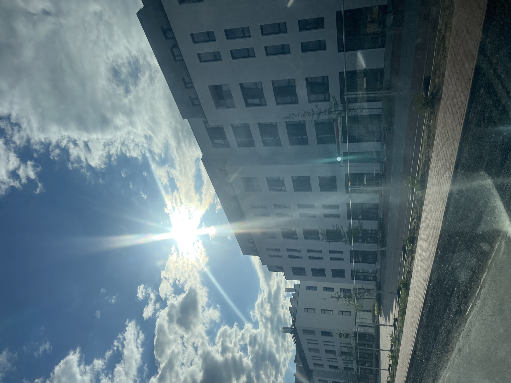 In Mynai, Ukraine, a new multi-story apartment complex is under construction, bathed in the bright sunlight of a partly cloudy day. The buildings are modern, with white facades and numerous dark-framed windows. Fencing is visible around the construction site, indicating ongoing development. A paved walkway runs alongside the buildings, suggesting an area that will soon be accessible to residents. The sky is a clear blue, with fluffy white clouds and a prominent sunburst effect caused by the bright sun. The overall scene depicts urban development and a bright, sunny day.