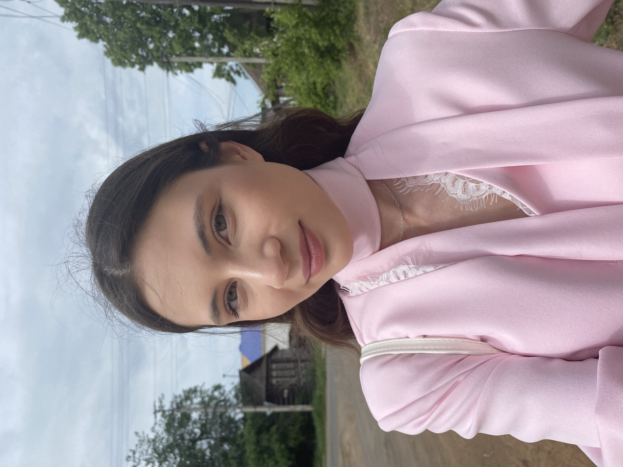 A woman with dark brown hair, light skin, and light brown eyes wearing a pink blazer with white lace trim is standing outside on a paved road. She is facing the camera and slightly smiling. In the background, there is a wooden structure with a blue and yellow tarp over it, green trees, and an overcast sky. This photo could have been taken in Votkinsk, Russia, based on the image context.