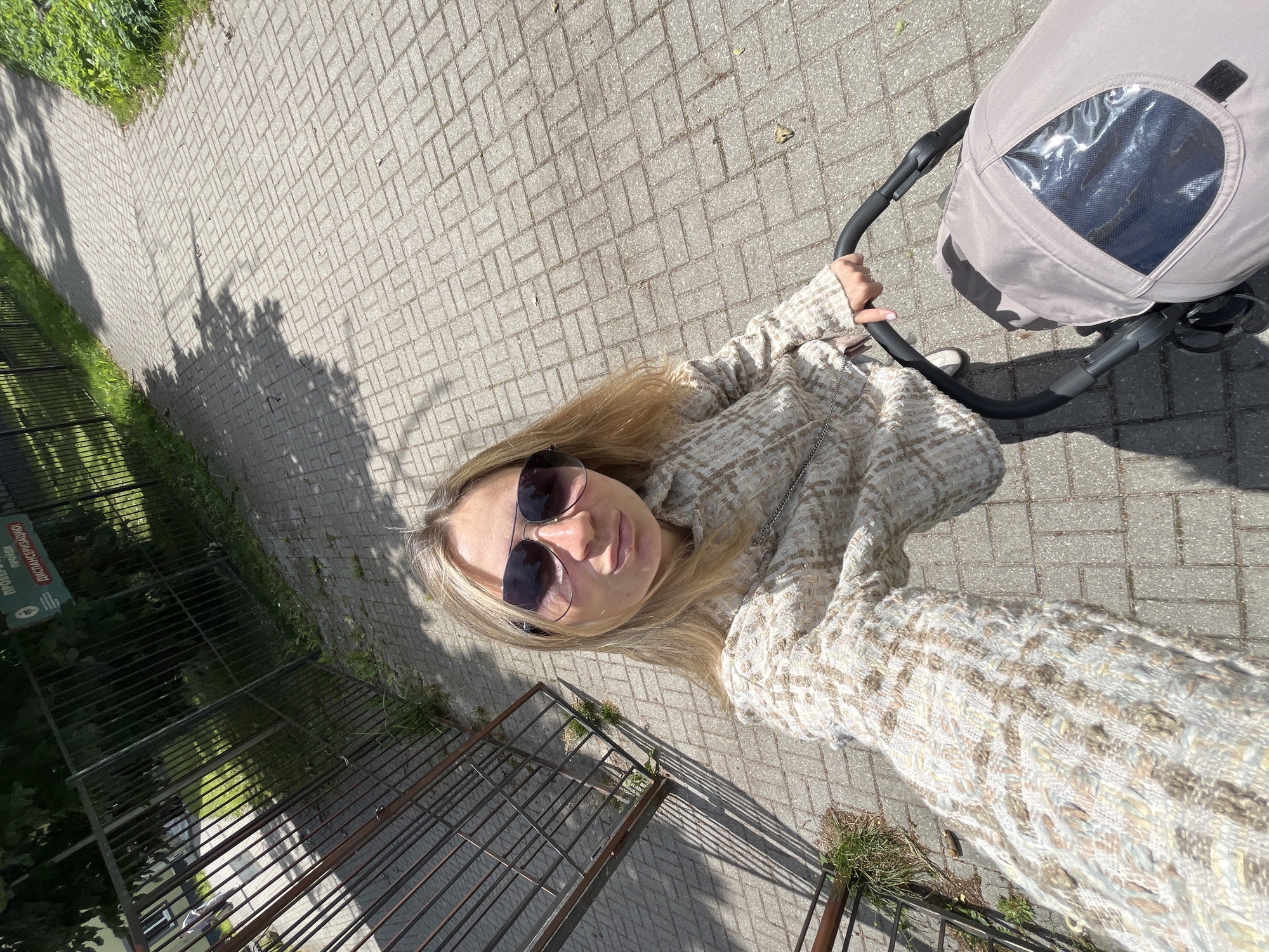 A woman with blonde hair and sunglasses is taking a selfie while pushing a stroller on a paved walkway. She is wearing a textured jacket. The walkway is bordered by a fence and grass on the left and more grass on the right. Tree shadows are cast on the walkway. A sign is visible on the fence. The location is Pionersky, Russia.
