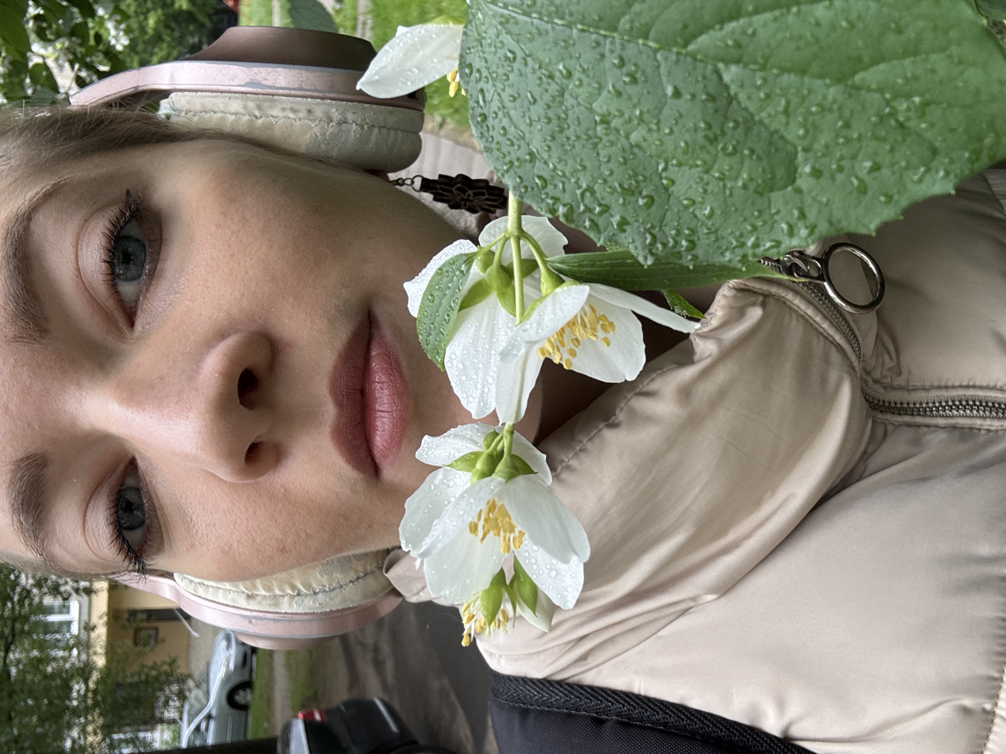 The image is a close-up, selfie-style shot of a woman wearing rose-gold headphones and a beige jacket. She is holding a sprig of white jasmine flowers with yellow centers, some of which are covered in water droplets. The flowers are in the foreground, obscuring part of her face and jacket. In the background, a building with yellow walls and a parked silver car are visible, suggesting an urban setting. The overall lighting indicates it is daytime, possibly overcast or after rain due to the water on the flowers and leaves. The woman's face is partially visible, with her blue eyes and pink lips showing. An earring with a dark, geometric design hangs from her right ear. The scene appears to be outdoors, on a street or sidewalk in the city of Minsk, Belarus.