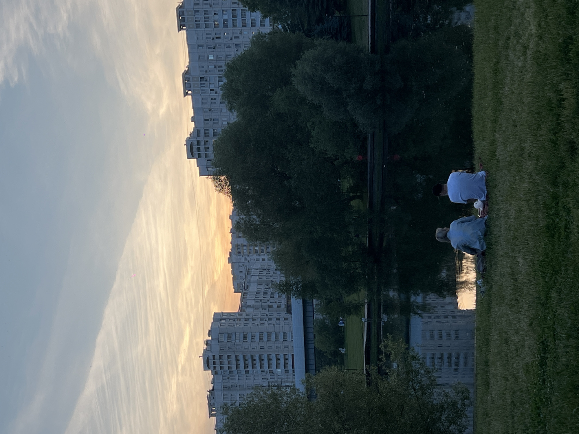 This image captures a dusk scene in a park situated within the city of Moscow, Russia, characterized by prominent residential architecture in the background. The sky, occupying the upper third of the frame, displays a gradient from light blue at the zenith to warm orange and pink hues near the horizon, indicative of sunset. Striated cirrus clouds are visible, with a distinct, elongated pattern across the upper-left sky. A small, bright pink luminous anomaly or object is present in the mid-upper-left section of the sky, near the cloud formation.
The background features numerous multi-story, grey residential buildings, typical of post-Soviet urban development, with visible windows and balconies. A dark, elevated bridge or overpass structure runs horizontally across the mid-ground, positioned between the buildings and the treeline.
The middle ground is dominated by a dense canopy of dark green trees, obscuring the lower portions of the distant buildings and the land immediately beyond the water. On the far side of the pond, a lighter green grassy bank is visible, with several small, indistinct figures appearing to be standing or walking.
The central and lower-middle section of the image is occupied by a calm, dark body of water, likely a pond or lake, which perfectly reflects the sky's warm colors and the distinct outlines of the distant residential buildings.
In the foreground, a verdant grassy area slopes gently towards the water. Two individuals are seated on the grass, facing away from the camera, towards the water and the sunset. The person on the left wears a light blue long-sleeved top and light-colored trousers, with light-colored hair. The person on the right wears a white short-sleeved t-shirt and light-colored trousers, with dark, short hair. They are positioned close together on a red blanket or mat. Near the person on the right, a white bag or container and a cup are visible on the mat. A dark object, possibly a bag, rests near the person on the left. The overall scene suggests a tranquil outdoor gathering during twilight.