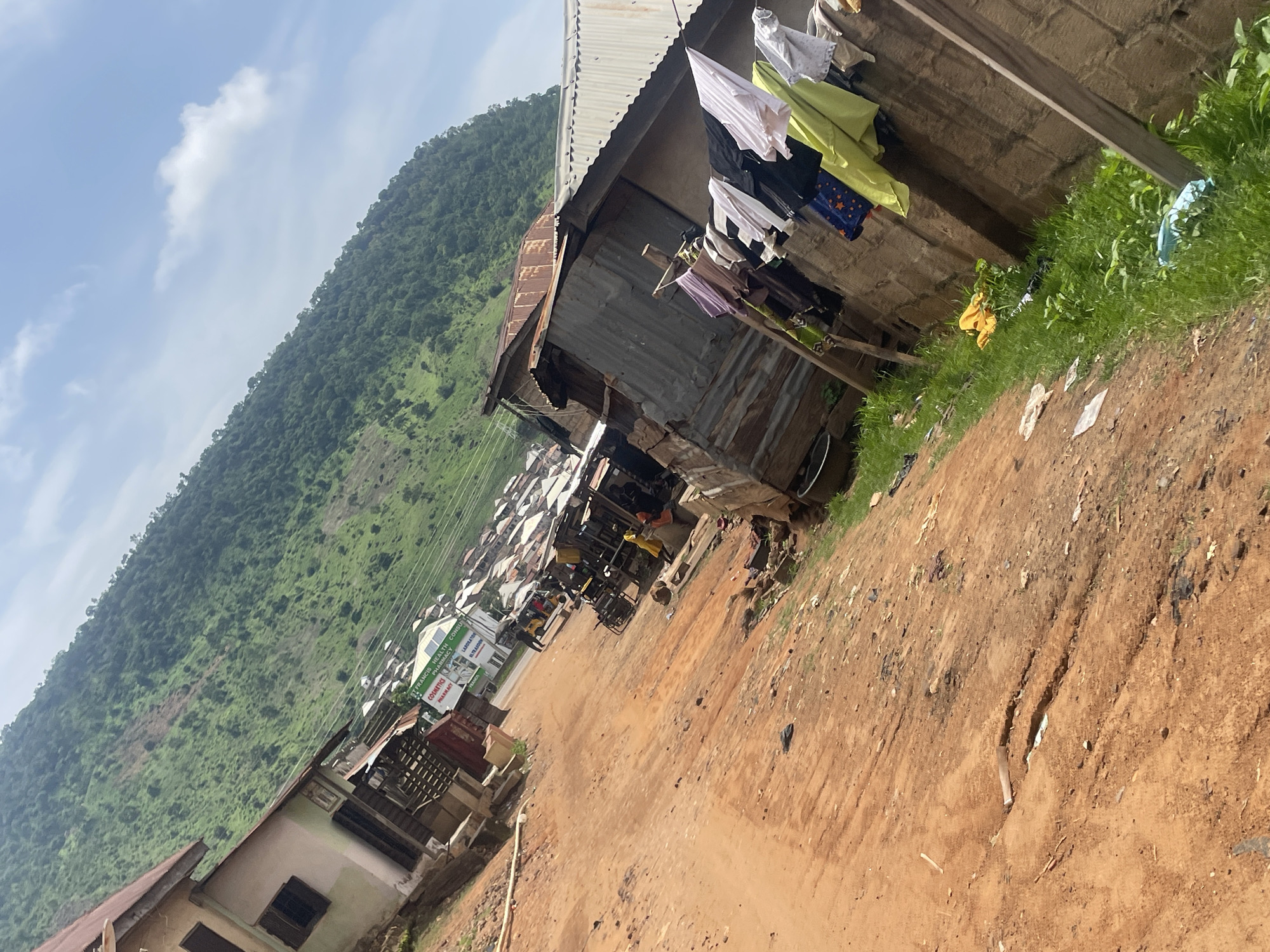 This image depicts a street scene in Lokoja, Nigeria, with a focus on a dirt road bordered by modest buildings and a lush, tree-covered hillside in the background. The road is uneven and appears to be unpaved, with some visible cracks and debris. On the right side of the road, a wall made of concrete blocks is visible, with laundry hanging on a clothesline stretched across it. The laundry consists of various articles of clothing in different colors, including white, black, purple, and bright green. Adjacent to the wall are several structures made of corrugated metal and wood, suggesting informal housing or small businesses. In the distance, a cluster of buildings with white roofs is visible, leading up to the steep, green hillside. A few people and vehicles, including a tricycle, are present on the road, indicating some level of activity. The sky above is blue with scattered clouds. The overall impression is of a community situated in a hilly, rural or semi-urban environment.