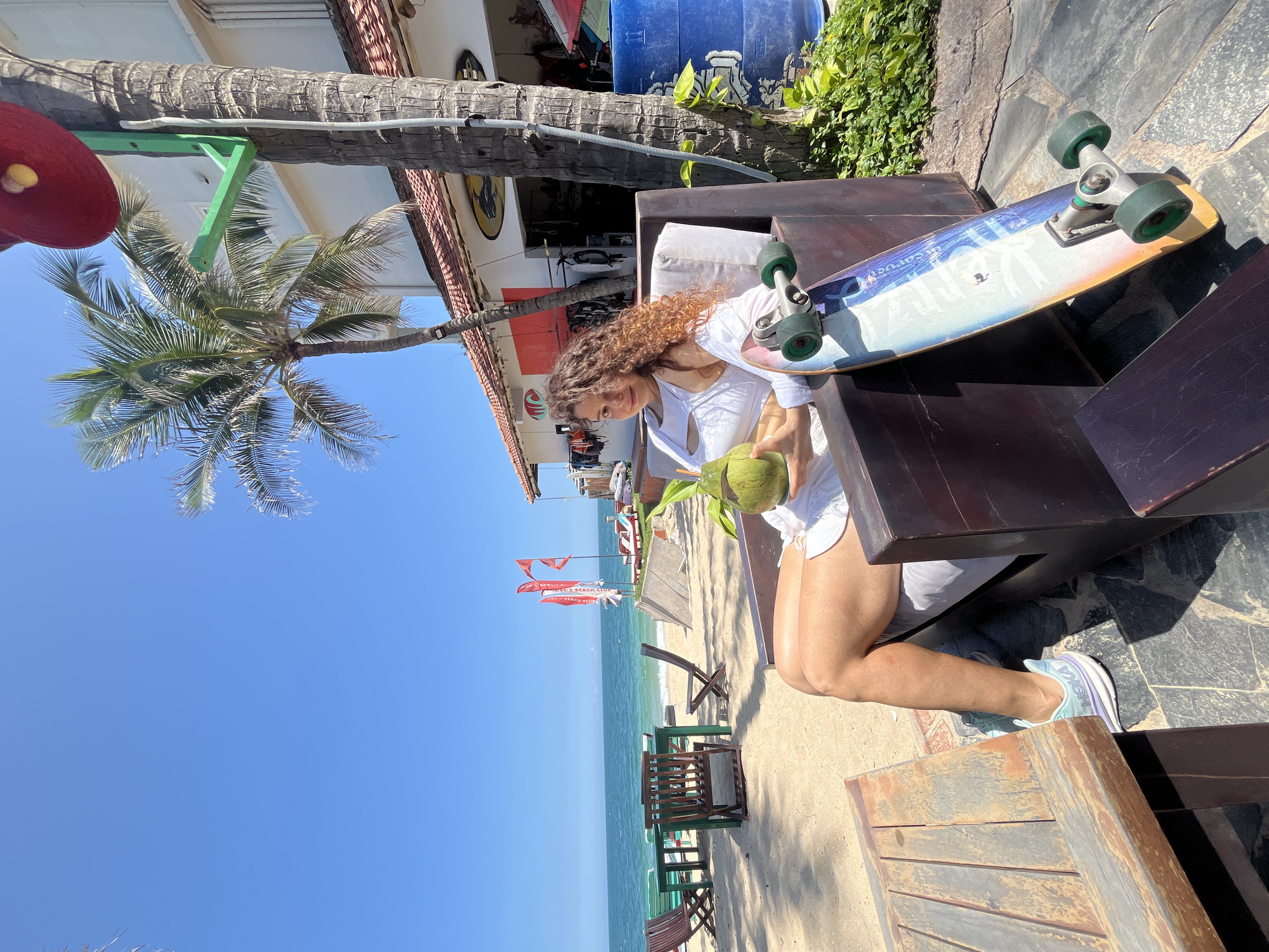 A woman with curly brown hair sits in a dark wooden chair on a sandy beach, holding a halved coconut with a straw. She is wearing a white crop top and white shorts. Beside her, leaning against the chair, is a skateboard with a colorful design. In the background, the turquoise ocean meets a clear blue sky. Beach chairs and tables are set up on the sand, and palm trees sway gently. A building with a tiled roof and windows is visible behind the woman, suggesting a beachfront establishment. The scene appears to be in Phan Thiết, Vietnam, a coastal city known for its beaches and water sports.