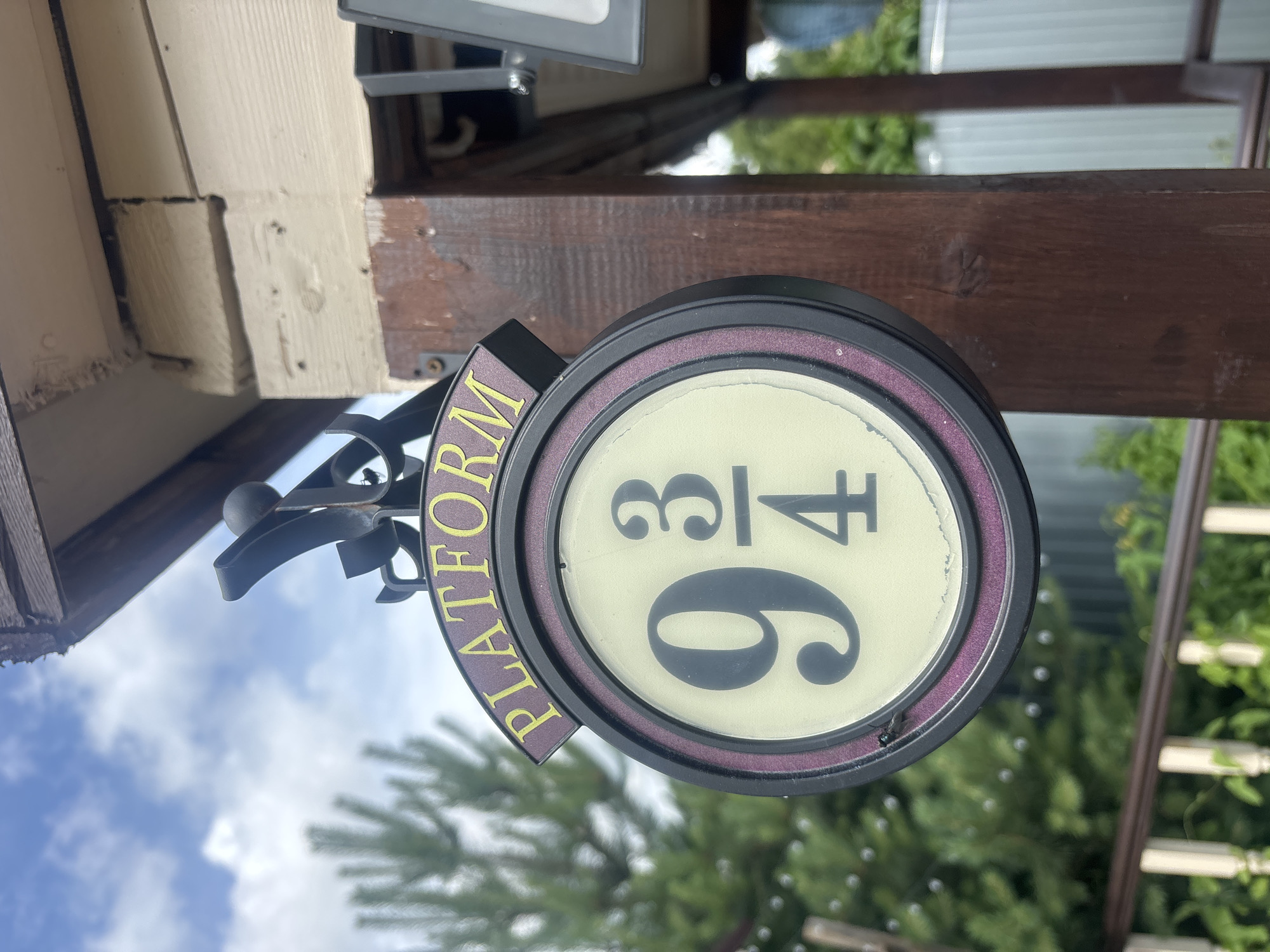 A close-up shot shows a sign for "Platform 9 3/4" hanging from a wooden post. The sign is round with a dark rim and a cream-colored center, featuring the number "9" in large font, and "3/4" in smaller font above and below a horizontal line. The word "PLATFORM" is written in yellow letters on a purple curved piece above the round sign. The background is slightly blurred, showing green trees and a blue sky with white clouds. The wooden post appears to be part of an outdoor structure.