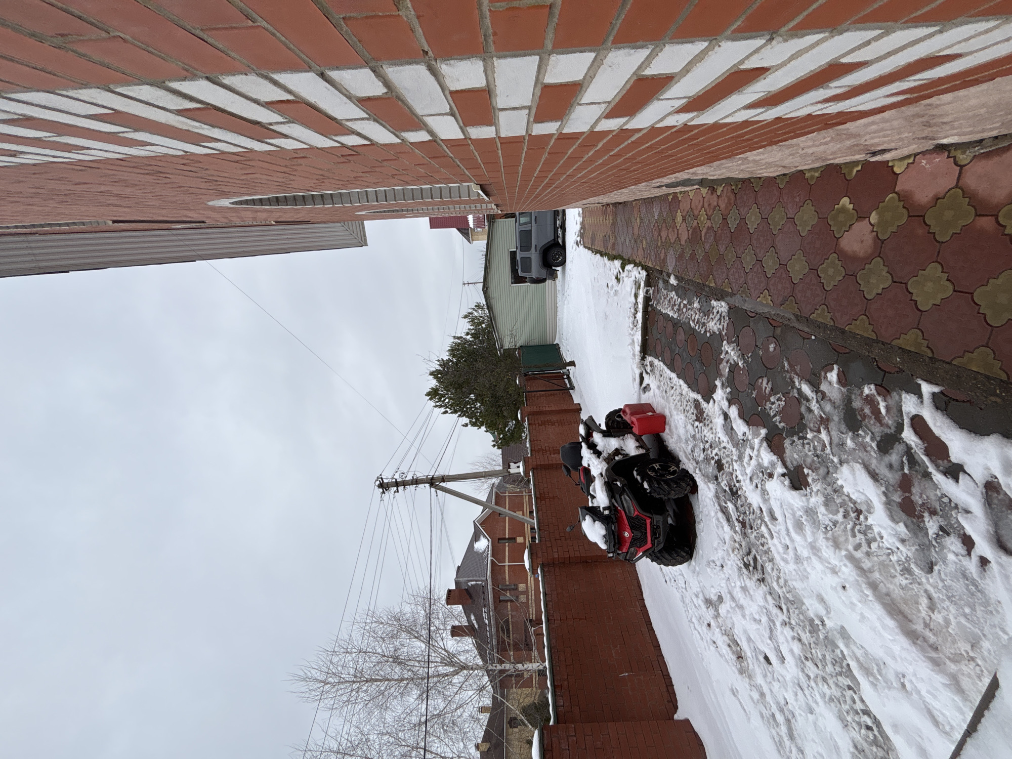 The image depicts a snowy alleyway between two brick buildings in Ферма, Russia. An all-terrain vehicle (ATV) is parked on the snow-covered walkway. The sky is overcast. Paved tiles form a decorative walkway in the alley. Overhead power lines are visible.