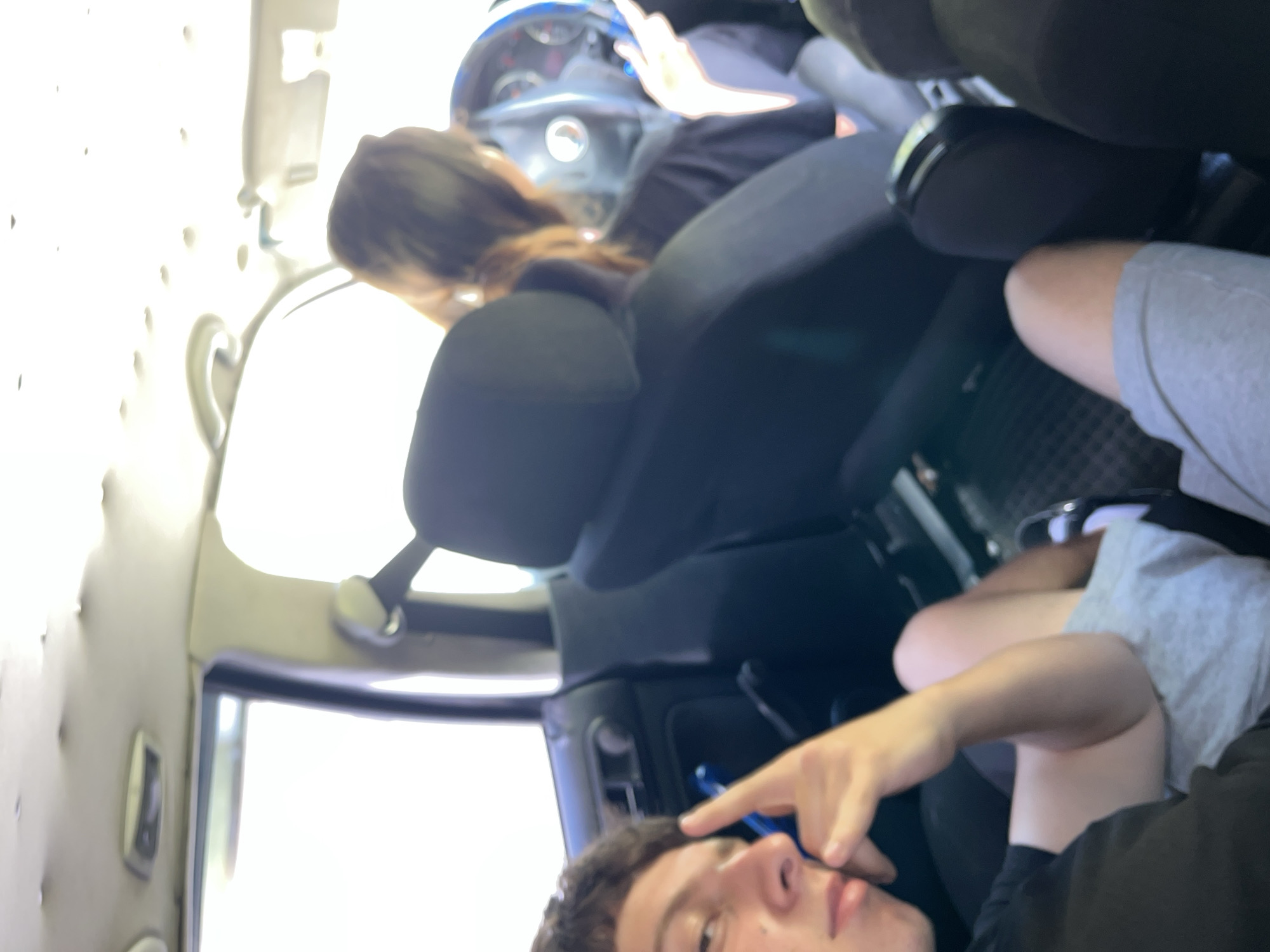The image depicts the interior of a car, likely in Vlahovo, Bulgaria. Two individuals are visible. In the foreground on the left, a young person with short dark hair is looking towards the camera, resting their chin on their hand and making a peace sign with their other hand. They are wearing a black t-shirt and light grey shorts. To the right and slightly behind, a person with long brown hair, presumably the driver, is looking forward and appears to be holding the steering wheel. They are wearing a black t-shirt. The car's interior shows black seats and a dark dashboard with a steering wheel. The headliner of the car appears to be damaged or sagging in places. The bright exterior light suggests it is daytime.