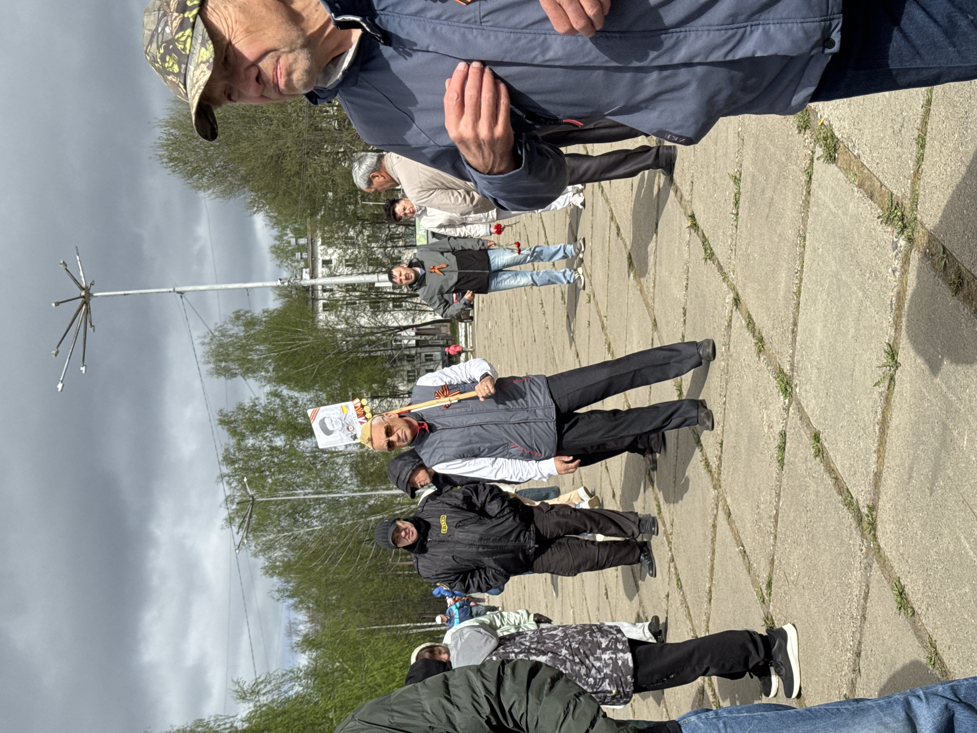 A group of people stand on a concrete paved area with grass growing in the seams, possibly in the city of Chaikovsky, Russia. The sky is overcast. A man in a grey jacket and black trousers holds a sign with a portrait of a person. Some people hold red flowers. Several other people are standing in the background. In the background are leafless trees, buildings, and a tall metal structure with radiating arms. A man wearing a camouflage hat stands in the foreground with his hand raised. Most people are dressed in casual clothing.