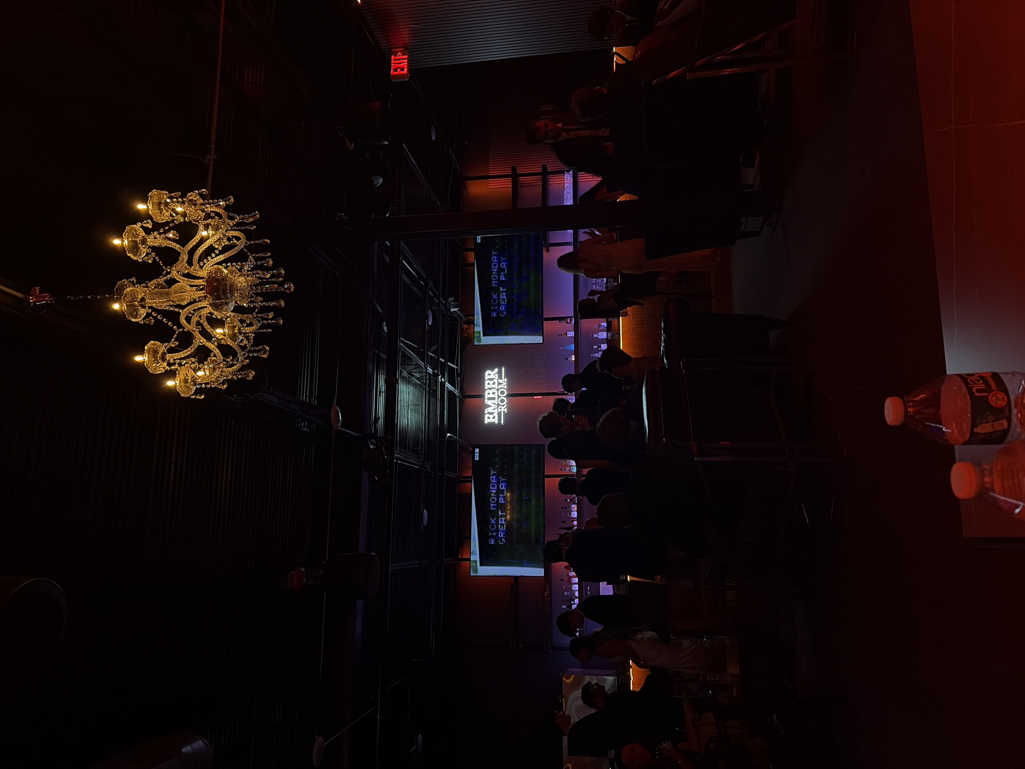 The image depicts the interior of "Ember Room," likely a bar or lounge, featuring a chandelier and a television screen displaying the phrase "RICK MONDAY. GREAT PLAY." Patrons are seated at a bar with alcohol and other drinks behind them. The venue appears to be dimly lit with a modern aesthetic, and the overall atmosphere suggests a social or entertainment setting. The image was likely taken in San Antonio, United States of America.