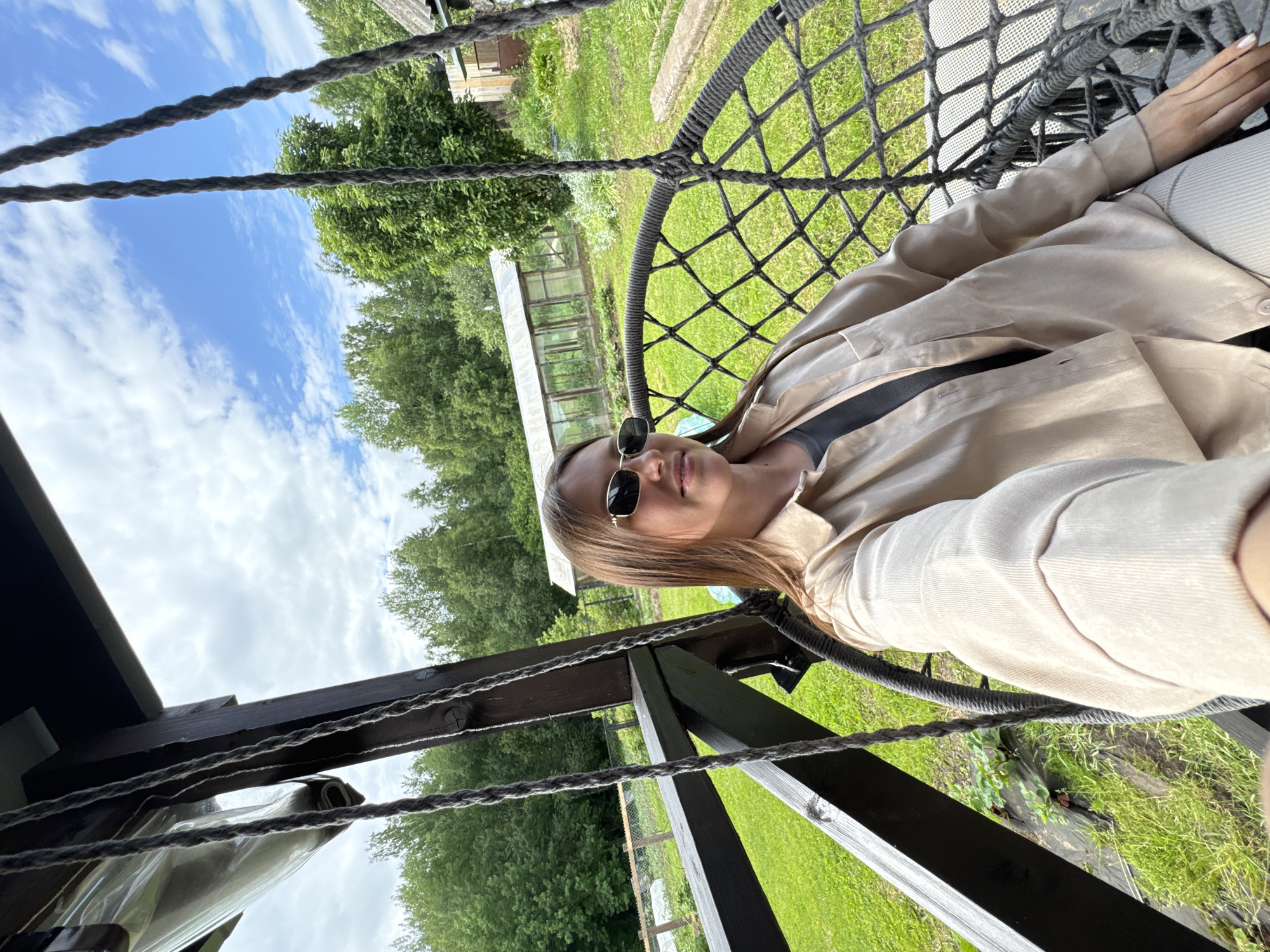 A young woman with shoulder-length brown hair and sunglasses is taking a selfie while sitting in a hanging chair. She is wearing a beige collared shirt and light-colored pants. The hanging chair is made of dark rope netting and is suspended from a wooden structure. The background shows a lush green yard with trees, a greenhouse, and a rural house. The sky is blue with scattered clouds. The image appears to be taken in the city of Ponaryevo, Russia, likely in a residential or recreational outdoor setting.