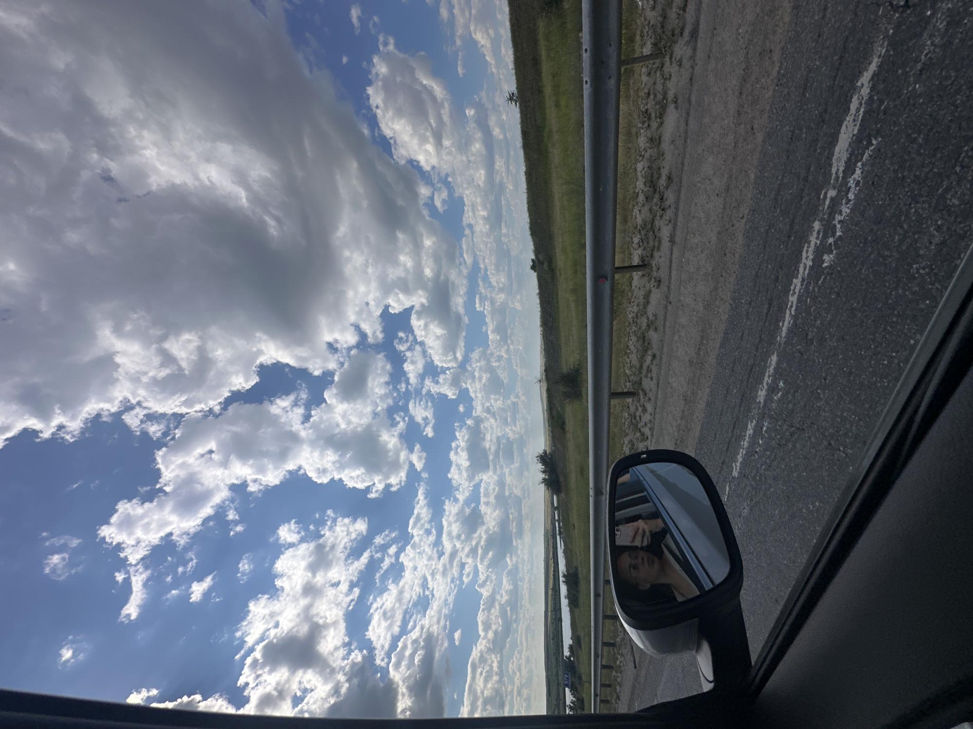 The image shows a view from a car window looking out at a landscape with a cloudy sky. In the side mirror, a young woman is visible, holding a phone and taking a selfie. The road is visible in the foreground, with a metal guardrail separating it from a grassy embankment and a body of water in the distance. The sky is dominated by large, fluffy white clouds against a blue backdrop. The location is identified as Ferma Sovkhoz Balykleyskiy, Russia.