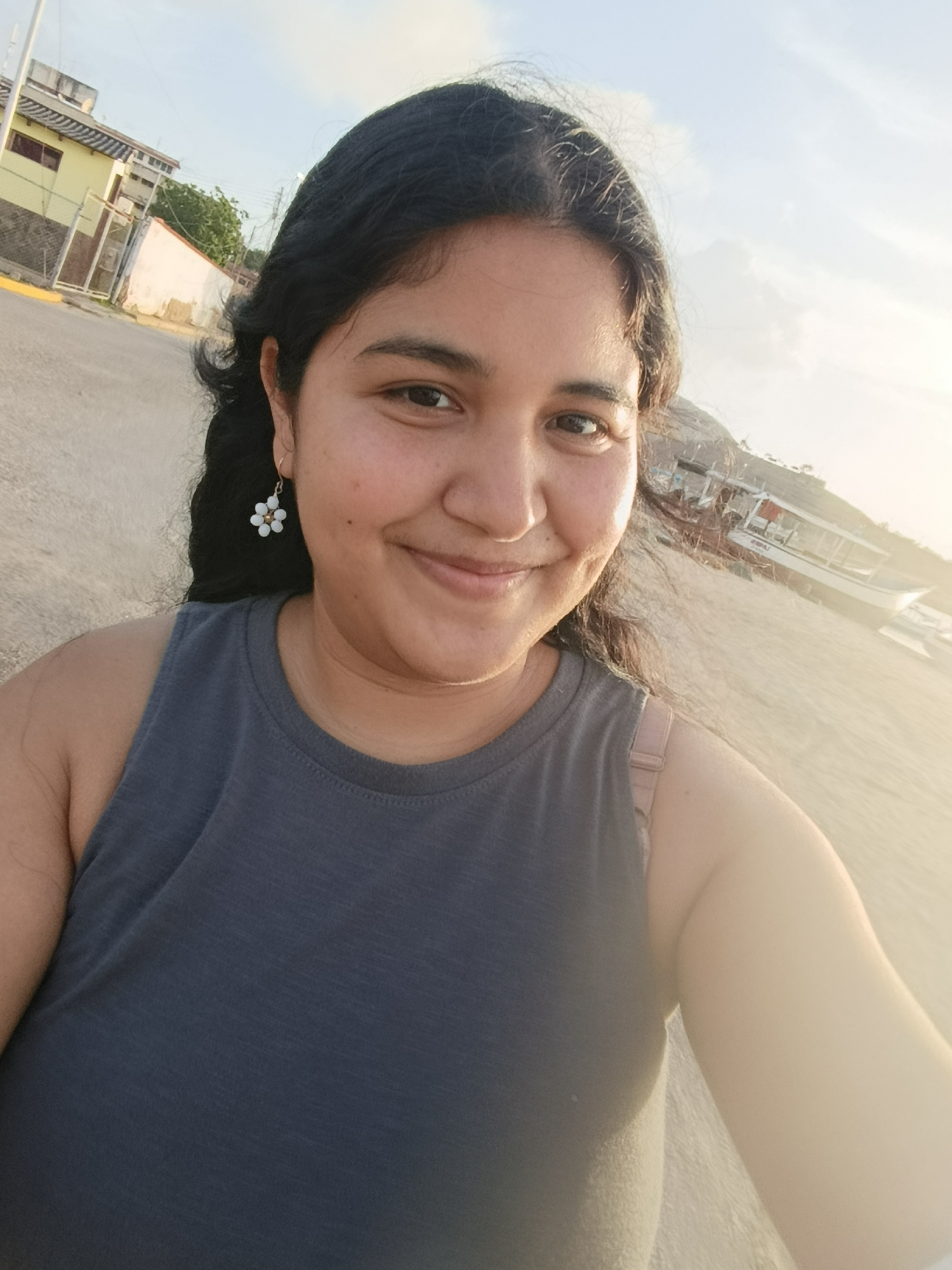 A young woman with dark hair and a grey tank top smiles at the camera outdoors. She is wearing a flower-shaped earring on her left ear. The background shows a sandy area with a white boat and some buildings in the distance. The lighting suggests it is late afternoon or early evening. The scene is likely in the vicinity of Juan Griego, Venezuela, given the presence of boats and a coastal environment.