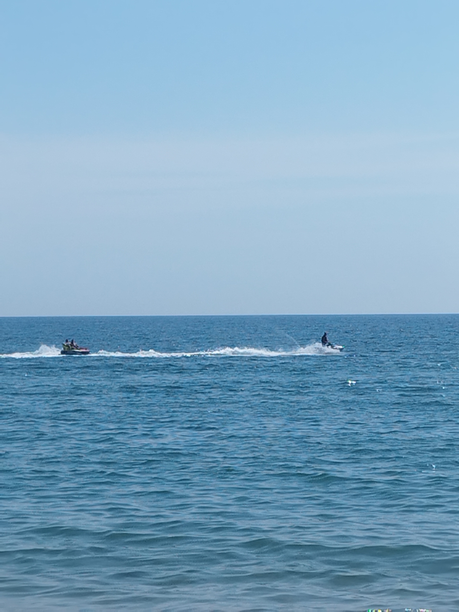 The image depicts a vast expanse of calm, blue ocean under a clear sky. The horizon line is sharp and separates the water from the sky. On the left side of the image, a small boat with two people is moving, leaving a white wake behind it. On the right side, a person is riding a jet ski, also creating a significant wake. Both watercraft are relatively small in the frame, emphasizing the immense scale of the ocean. The water is a deep blue, with gentle waves visible throughout. The scene appears to be a sunny day, likely at a beach or coastal area. Given the location of Constanța, Romania, this image likely captures a scene along the Black Sea coast.