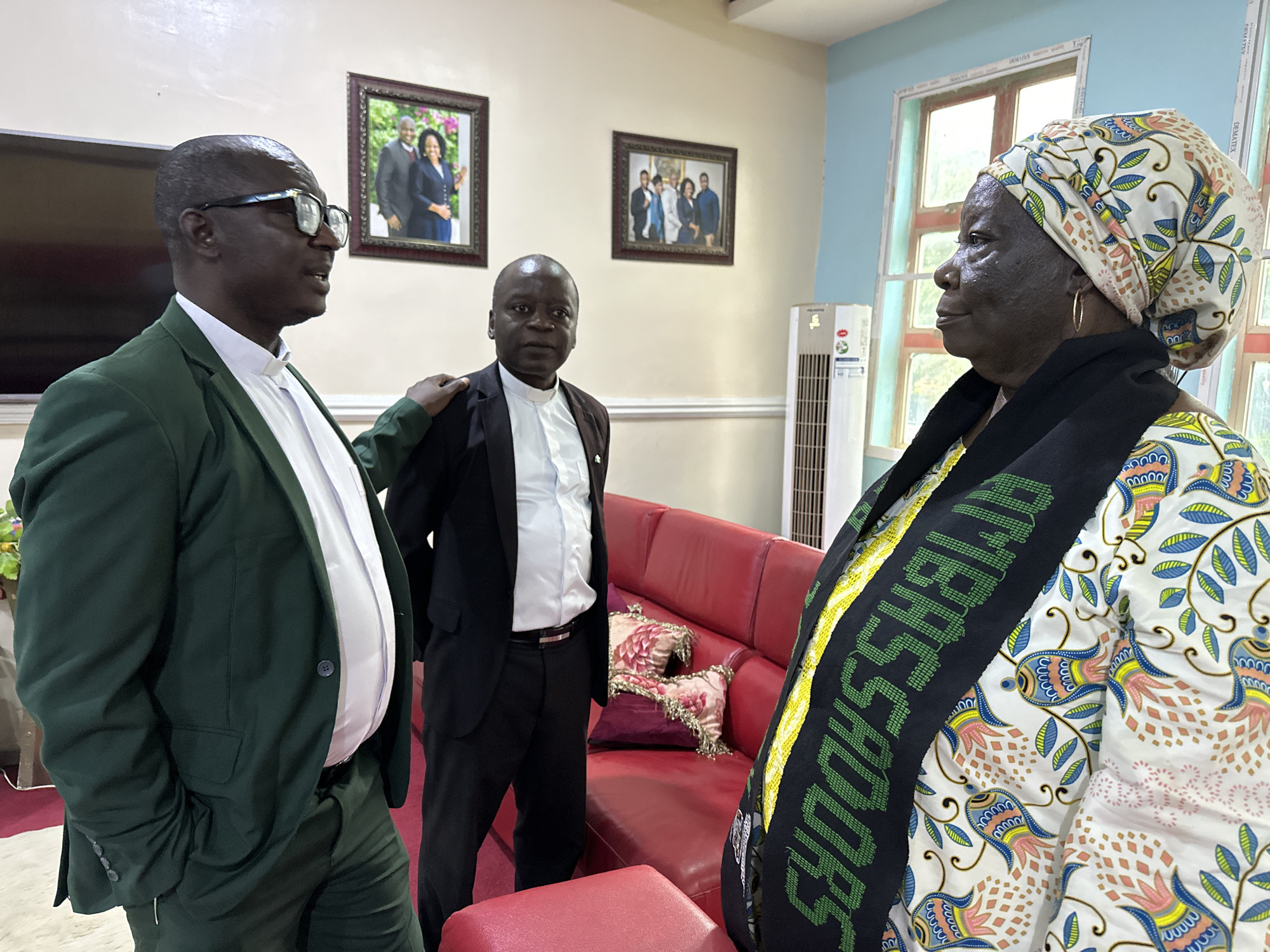 In Television, Nigeria, three individuals are gathered indoors, possibly in a home or office setting, evidenced by the presence of framed photographs on the wall and a red sofa. A man on the left, wearing a green suit and glasses, engages in conversation with another man in a black suit and white shirt, who stands to his right. A woman in a colorful headwrap and patterned attire is positioned on the far right, also appearing to be part of the conversation. Behind them, a tall white air conditioning unit stands against a light blue wall. The room is decorated with two framed pictures on the wall depicting groups of people. The overall atmosphere suggests a meeting or social gathering.