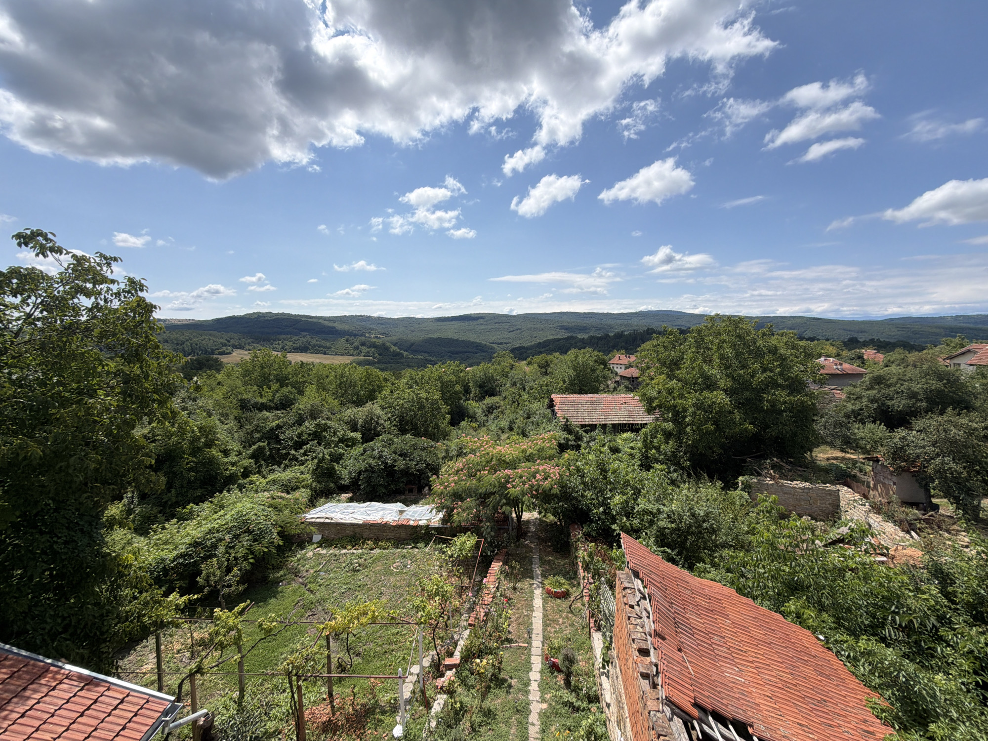 This is a high-angle, wide shot photograph of a rural landscape in Gorno Pavlikene, Bulgaria, on a partly cloudy day. In the foreground, the red-tiled roof of a building is visible, extending from the bottom left to the bottom right of the frame. Beyond this, a courtyard with a narrow stone pathway is seen, flanked by green grass and low-lying vegetation. A chain-link fence runs along the left side of the courtyard, with a trellised vine growing on it.
Further into the scene, there are remnants of old buildings, including a structure with a white, translucent roof and a partially collapsed stone wall. A tree with pink blossoms stands prominently in the middle ground, surrounded by dense green foliage. In the background, rolling hills covered in lush green trees stretch towards the horizon, under a bright blue sky with scattered white and grey clouds. The overall impression is one of a rustic, slightly overgrown village setting.