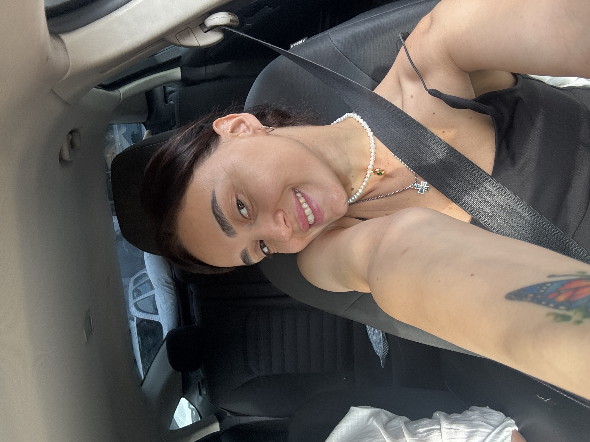 A woman is taking a selfie in the passenger seat of a car. She is wearing a black tank top and a pearl necklace with a gold charm. A seatbelt is visible across her body. Her arm, with a butterfly tattoo, is resting on her shoulder. The background shows the interior of the car and parked cars outside the window. The scene is not identifiable as being in Tbilisi, Georgia.