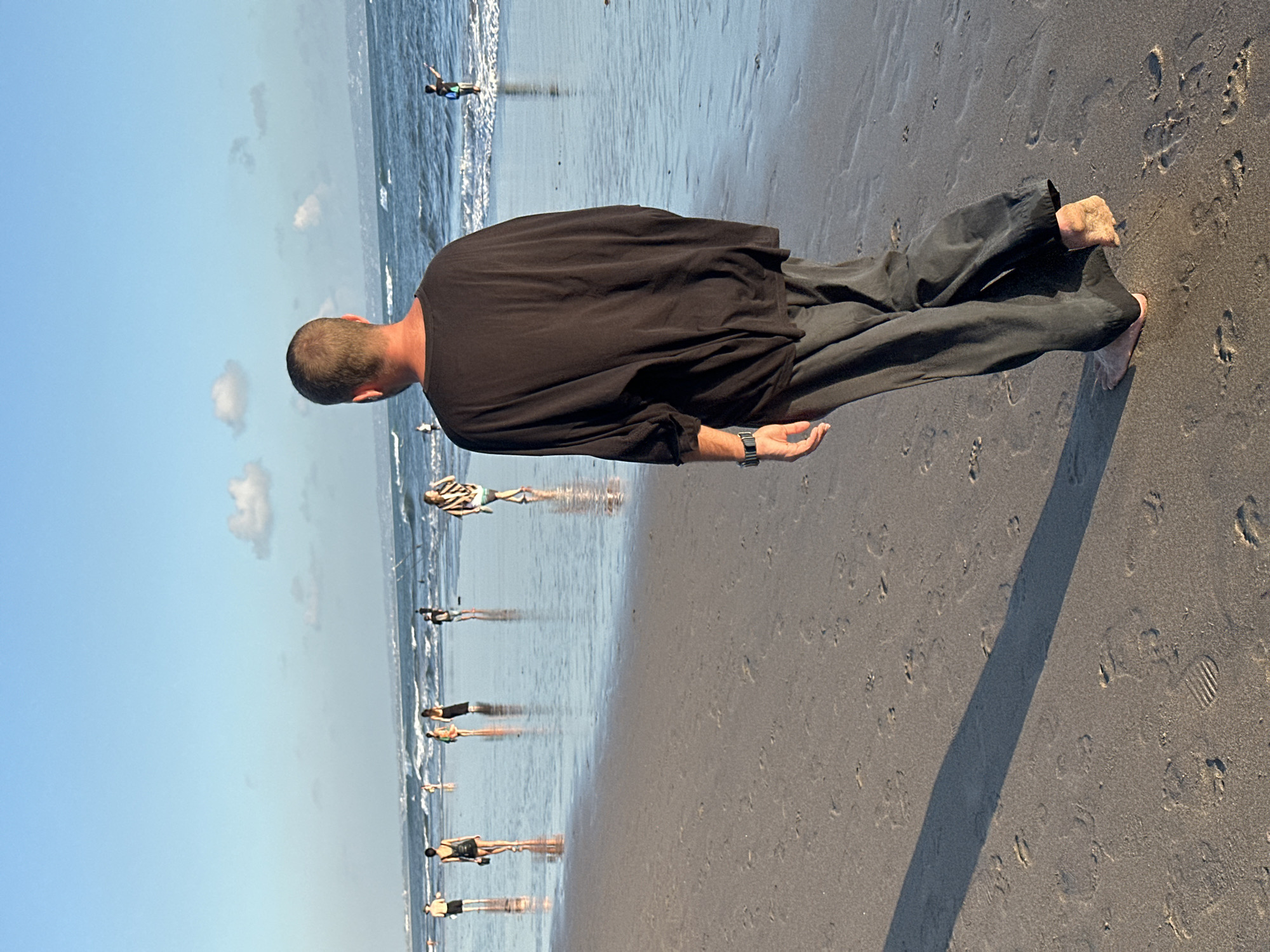 A man walks barefoot on a wet sandy beach in North Kuta, Indonesia. He is wearing a black, oversized, short-sleeved t-shirt and dark grey wide-leg pants. The man is seen from behind, facing the ocean. Other people are scattered on the beach and in the shallow water. The sky is clear with a few scattered clouds, and the sun is low in the sky, casting long shadows. The waves are moderate, and the water appears calm. The beach is relatively wide and flat, with footprints visible in the sand. The overall scene depicts a relaxed and peaceful atmosphere.