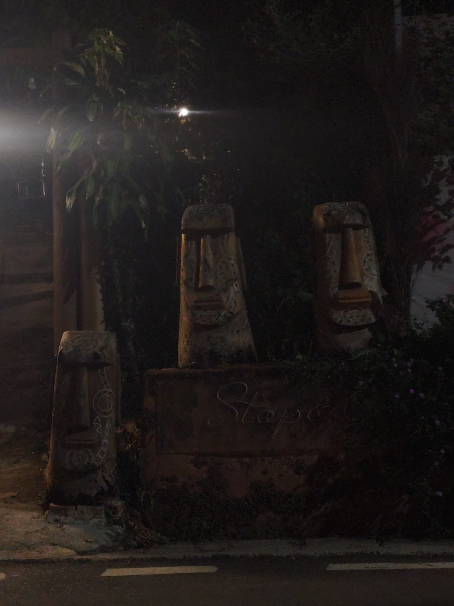 The image shows three statues resembling Easter Island heads at night. The statues are arranged on a platform with the word "Stop" inscribed on it. Dense foliage surrounds the statues, and a street with white lane markings is visible in the foreground. The location is possibly Da Lat, Vietnam, based on the style of the statues and general atmosphere. No people are visible in the image. A street light is seen illuminating the statues, with some additional light appearing on the left side of the image.