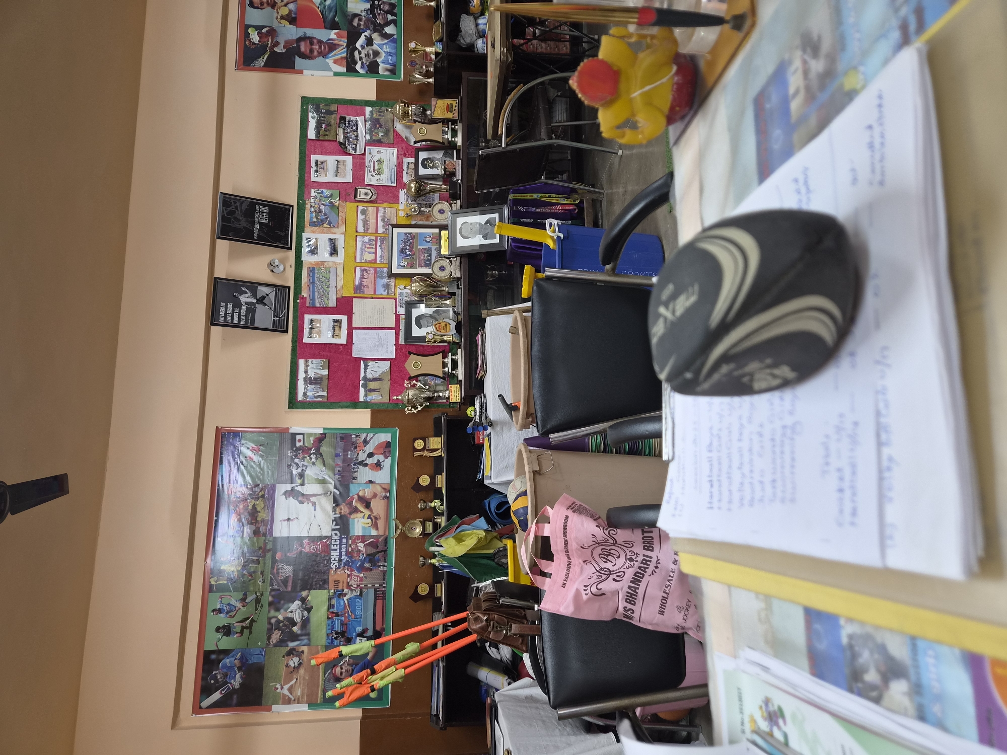 The image depicts an indoor room, possibly a sports office or equipment storage area. The walls are adorned with numerous photographs and posters showcasing various sports, athletes, and possibly past sporting events. There are multiple shelves filled with trophies, awards, and sports equipment. In the foreground, there are desks with papers, a rugby ball-shaped mouse, and a pink shopping bag with text indicating it's from "N/S BHANDARI BROTHERS WHOLESALE & RETAIL". Various chairs are visible, some occupied by sports equipment like hurdles. A ceiling fan is visible at the top of the frame. The overall impression is one of an organized, yet busy, space dedicated to sports. The mention of "N/S Bhandari Brothers Wholesale & Retail" suggests a commercial establishment, possibly a sporting goods store or a sports club office within Pathankot, India.