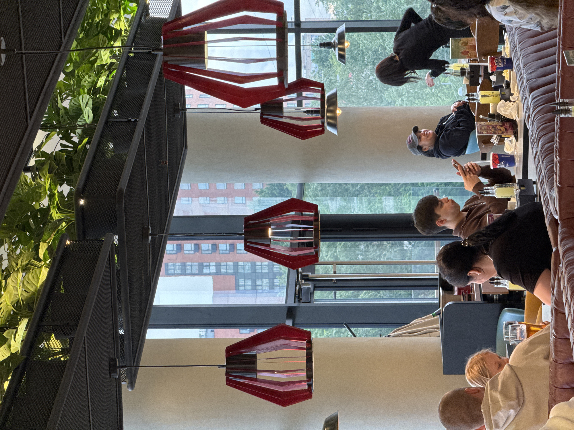 Here is a description of the image:
The image shows the interior of a restaurant, likely in the city of Moscow, Russia, based on the architectural style of the buildings visible through the windows. A group of people are seated at a long table. The people appear to be having drinks and conversations. The table is laden with various beverages and glasses. Above, distinctive red geometric pendant lights hang from the ceiling. A metal structure with plants is positioned on the upper left side of the frame. Large windows reveal buildings and trees outside.