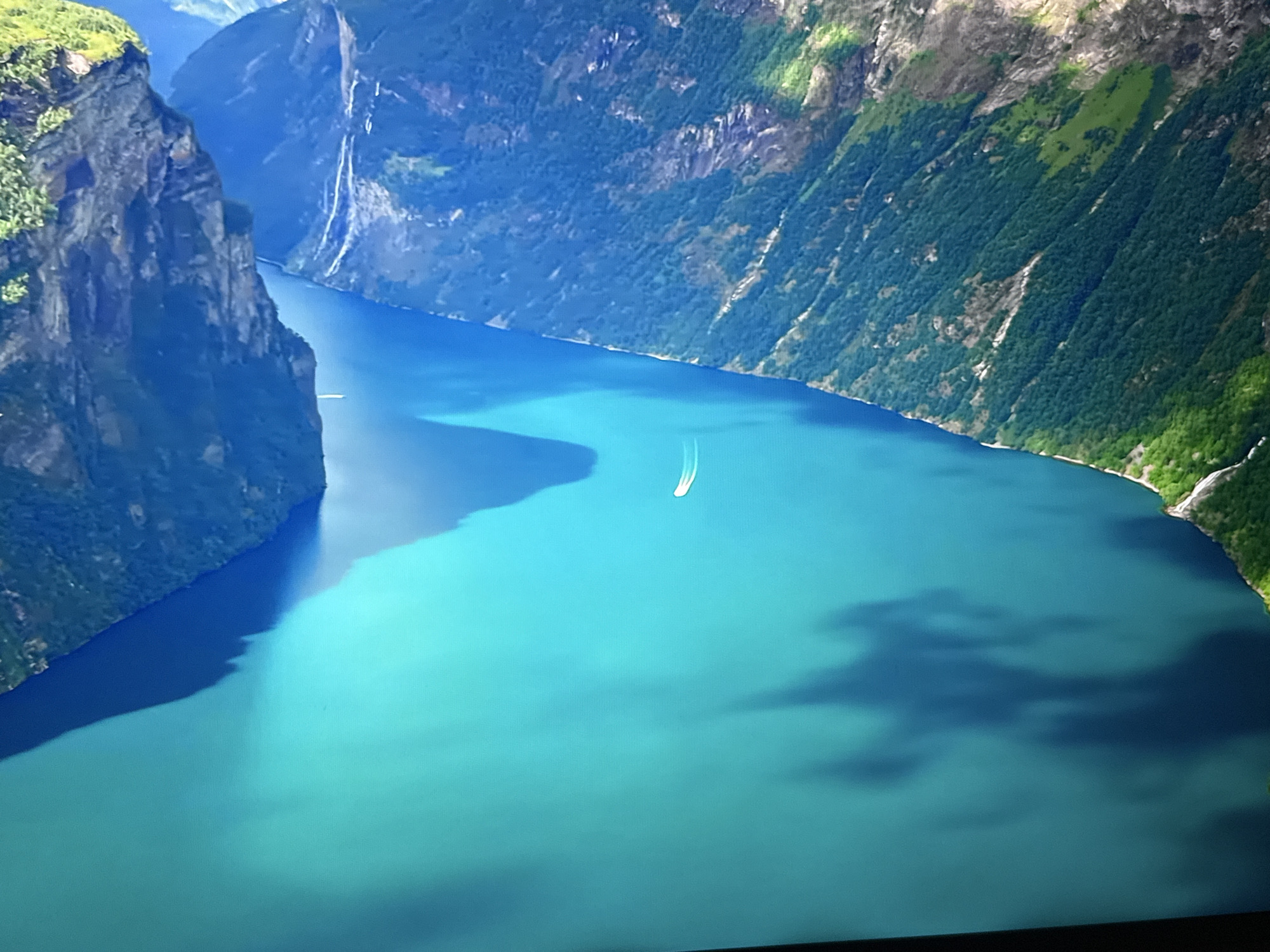 The image depicts a wide, tranquil fjord with steep, verdant cliffs on either side. The water is a striking turquoise color, reflecting the bright sky and the surrounding mountains. A small white boat is visible in the distance, leaving a faint wake on the water's surface. The cliffs are densely covered with lush green vegetation, interspersed with patches of rock. Shadows cast by the mountains create darker areas on the water, adding depth and contrast to the scene. The overall impression is one of natural beauty and serene remoteness. This image is not located in the city of Burgas, Bulgaria.