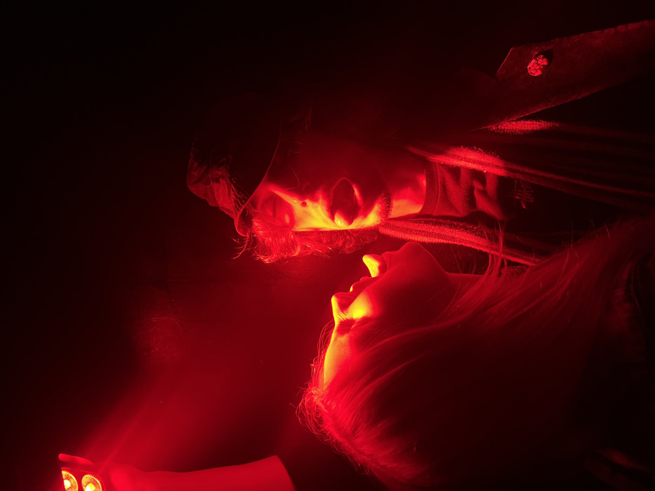 The image depicts an indoor scene in Paris, France, illuminated by intense red light emanating from a handheld device visible in the upper left. Two individuals are shown in close profile, facing each other. On the left, a female-presenting individual with long hair has her mouth open, angled upwards. On the right, a male-presenting individual wears a dark bucket hat, a patterned scarf, and a dark t-shirt with a white logo. His mouth is wide open, with the tongue extended. A small, possibly skull-shaped, pin is affixed to his outer garment near the left shoulder. The subjects' faces are brightly highlighted by the red light, suggesting an energetic vocal interaction like singing or shouting in a dimly lit environment such as a nightclub or music venue.