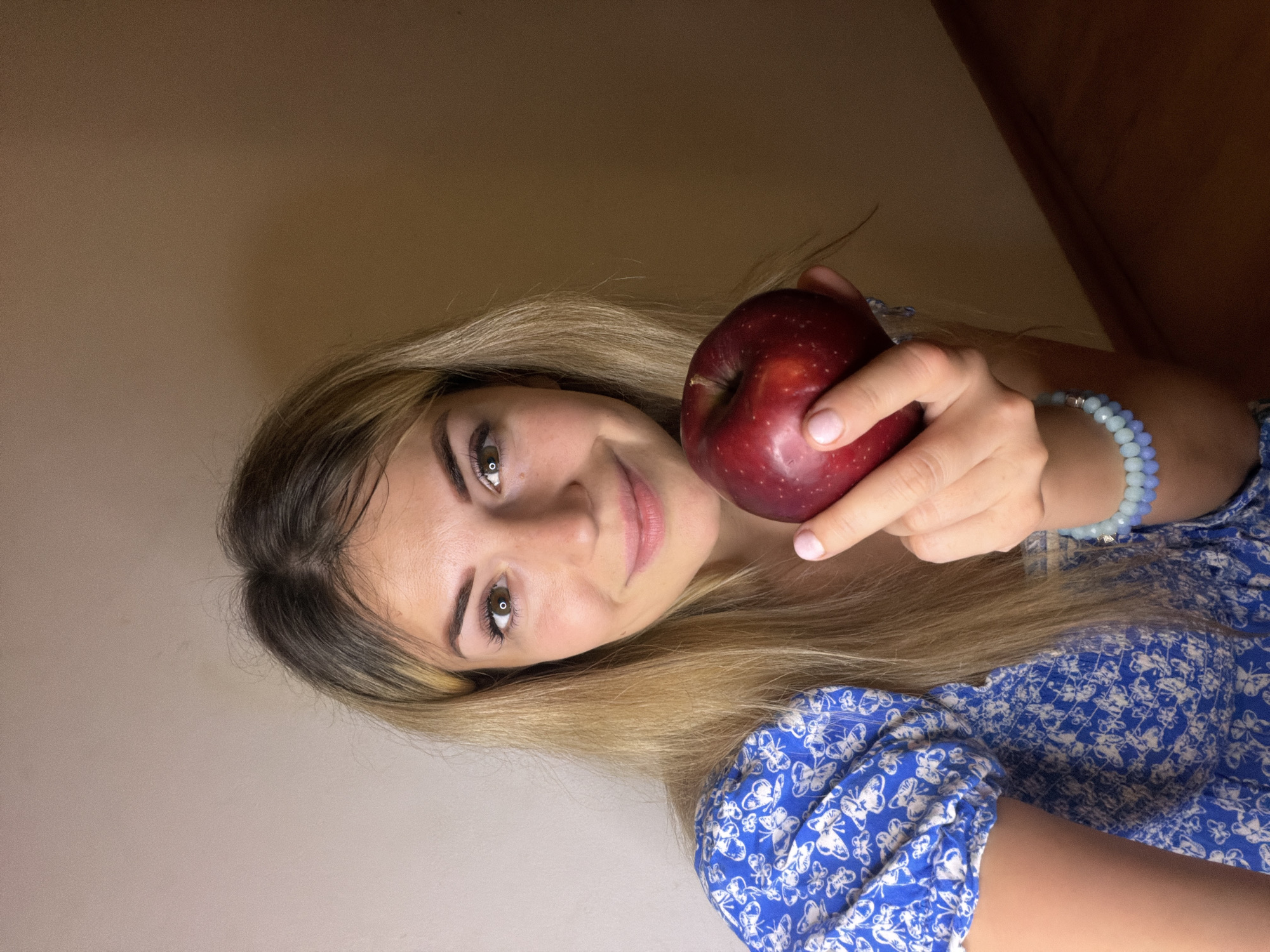 This is a portrait photograph featuring a young woman holding a red apple, taken in Budva, Montenegro. The woman is positioned centrally, looking directly at the camera with a gentle smile. She has long, straight hair with blonde highlights and darker roots, which frames her face. Her eyes are brown, and she wears light makeup, including eyeliner and what appears to be a subtle pink lip color.
She is dressed in a casual blue top or dress adorned with a repeating white butterfly pattern. On her right wrist, she wears a light blue beaded bracelet. Her left hand holds a shiny, deep red apple near her face, with her fingernails painted a light pink. The background is a plain, light brown or beige wall, suggesting an indoor setting. There are no other objects or people visible, focusing all attention on the woman and the apple. The lighting appears soft and even, without harsh shadows, hinting at artificial illumination or indirect natural light. The overall mood conveyed is serene and pleasant. There is no visible text in the image.