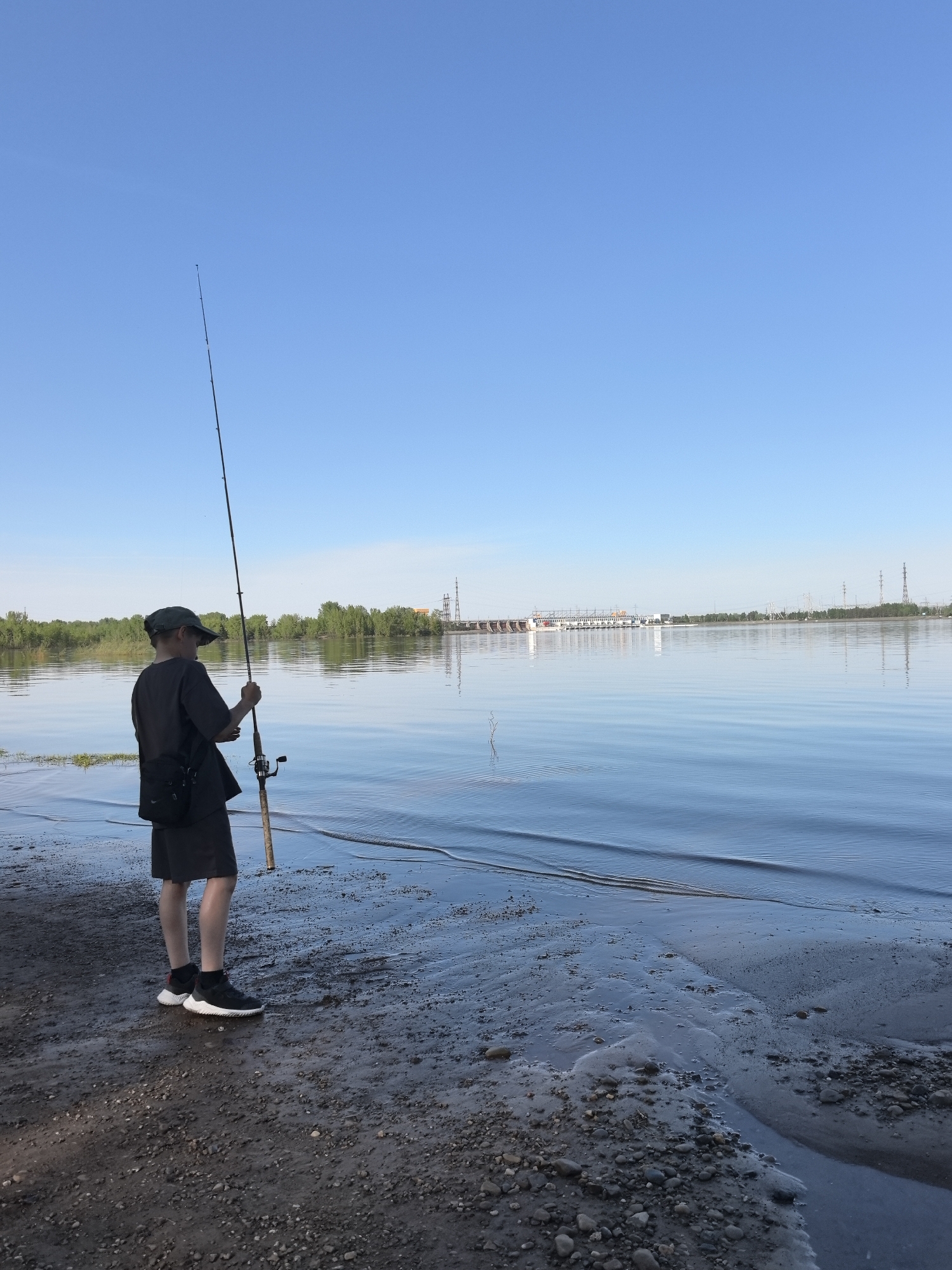 A young person stands on a wet, rocky beach, holding a fishing rod. The person wears dark clothing, including a hat, t-shirt, shorts, and athletic shoes. A dark bag hangs from their shoulder. The fishing rod is tall and slender, extending upwards into the clear blue sky. Behind the person is a wide expanse of water, with a structure resembling a dam or industrial complex visible on the opposite shore. Trees line the distant shore, and power lines are also visible. The scene is bathed in daylight, and the water is calm with small ripples. The location is near the city of Noviy, Russia.