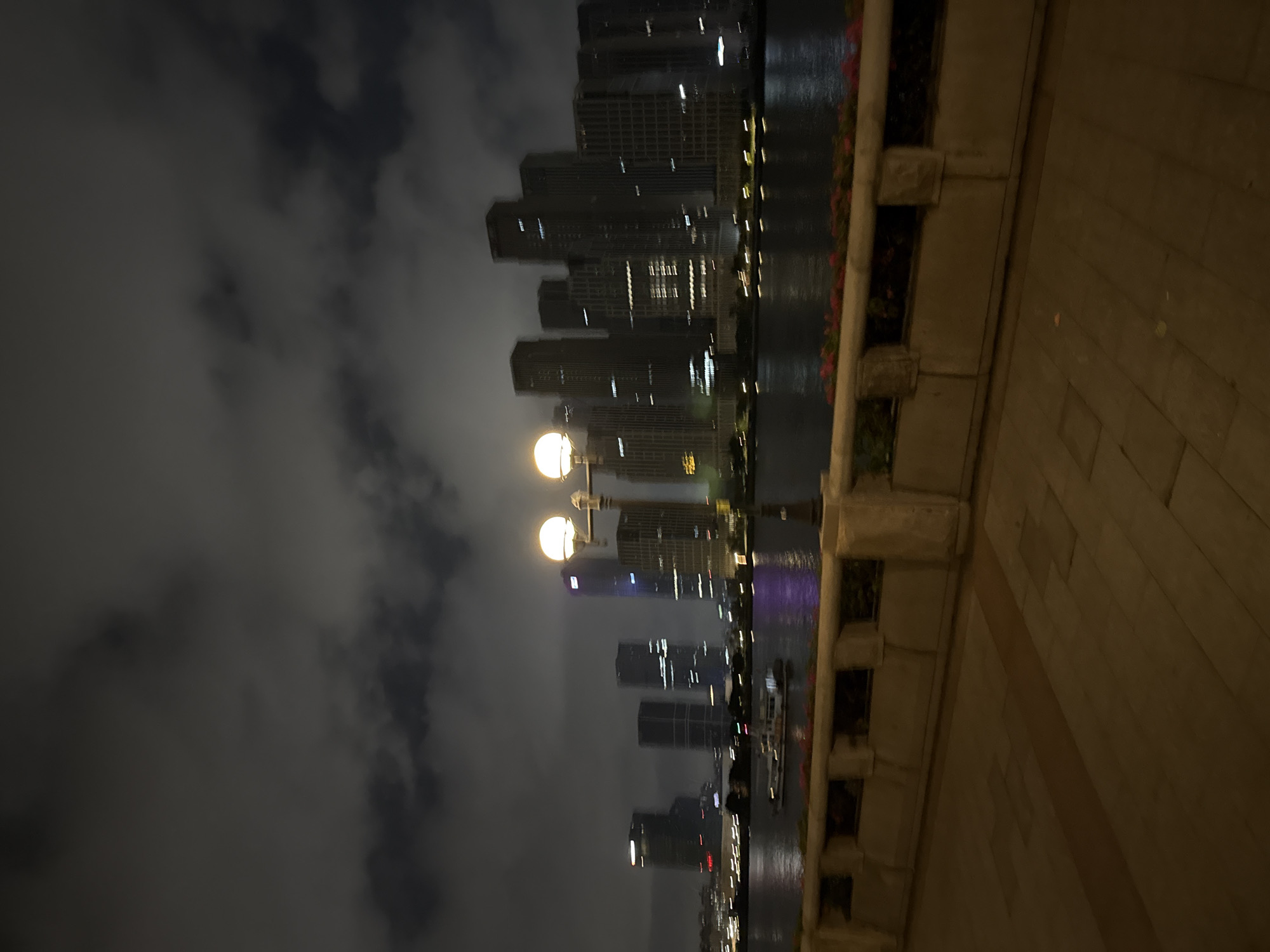 An exterior, nighttime, medium shot captures a waterfront scene in Haizhu District, China. A walkway with a stone railing and planters separates the viewer from a river or bay. In the distance, a dense cluster of modern skyscrapers with illuminated windows forms the city skyline. A boat is visible on the water, moving from left to right. A lamppost with two glowing spherical lights stands in the foreground, on the right side of the image, its light illuminating the walkway. The sky is cloudy and dark, typical of a night scene. The overall image has a slightly blurred quality, suggesting movement or a low-light condition.
