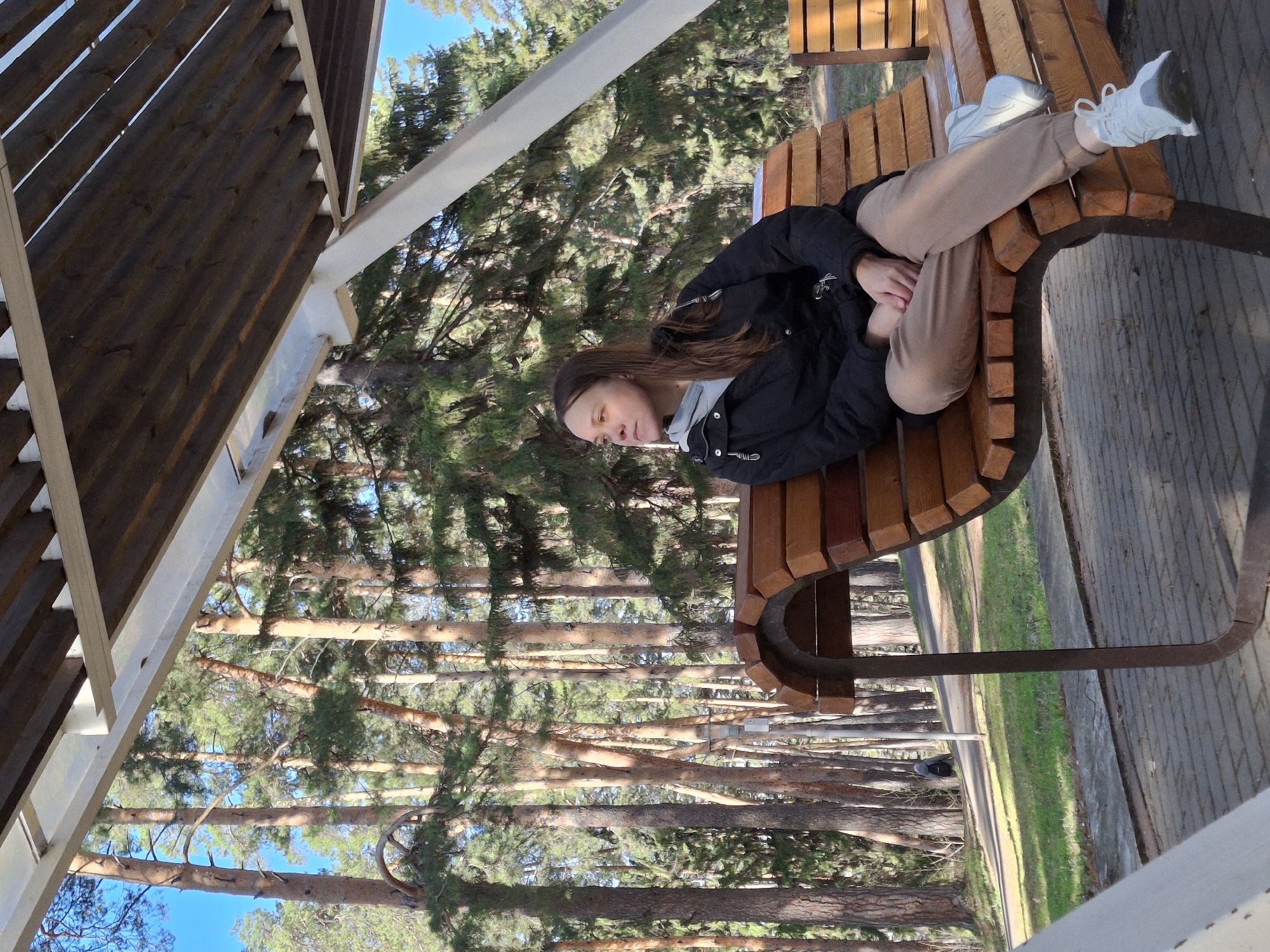 The image depicts a young woman relaxing on a curved wooden bench in an outdoor setting. She is lying on her back with her head towards the left side of the frame and her feet, clad in light-colored sneakers, crossed towards the right. Her arms are folded over her chest. She is dressed in a black puffy jacket over a light grey hooded top, and light brown or beige sweatpants. Her long, dark brown hair is visible.
The bench is constructed from light-colored wooden slats supported by dark metal framing. Above and to the left, part of a wooden and light-colored metal shelter structure or pergola frames the scene.
In the background, a dense forest of tall, slender pine trees dominates the landscape, with patches of clear blue sky visible through their canopies and above them. To the right of the bench, a paved pathway runs alongside grassy areas, receding into the distance.
The bright sunlight suggests a clear, pleasant day. The overall atmosphere conveys a sense of calm and relaxation, as the woman appears to be resting or contemplating. The setting is likely a park or forest recreation area in Sarapul, Russia. No visible text is discernible in the image.