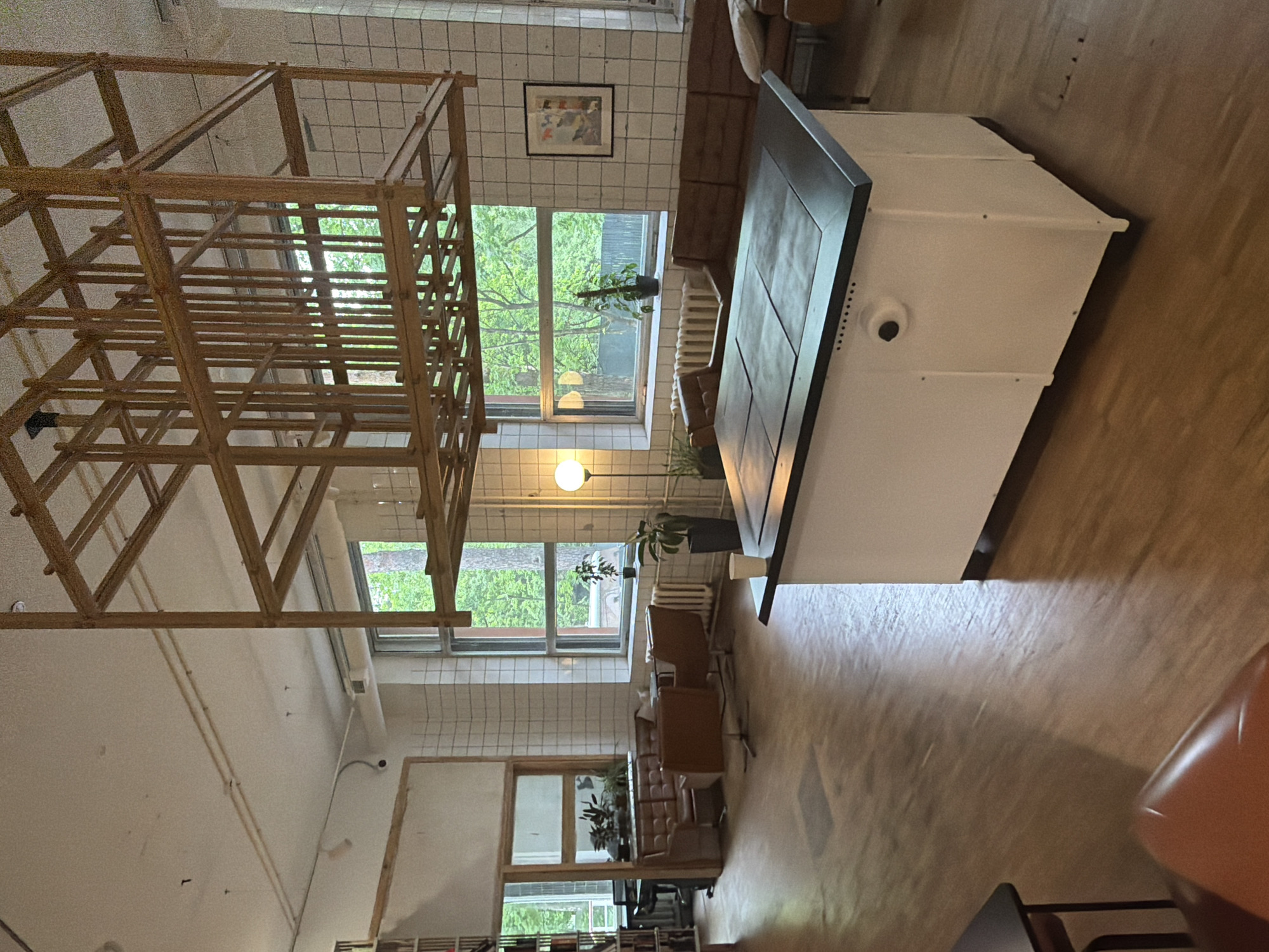 The image shows an interior space with a large wooden structure occupying a significant portion of the view, possibly an unfinished art installation or architectural element. The space features a tiled wall with two windows offering views of trees, suggesting a location within a building. A picture hangs on the tiled wall above one window. A white object with a dark top sits near the windows, possibly a kitchen appliance. A seating area is visible with chairs and a sofa-like structure. The overall environment is well-lit with natural light coming through the windows. No people are discernible. The location is unknown.