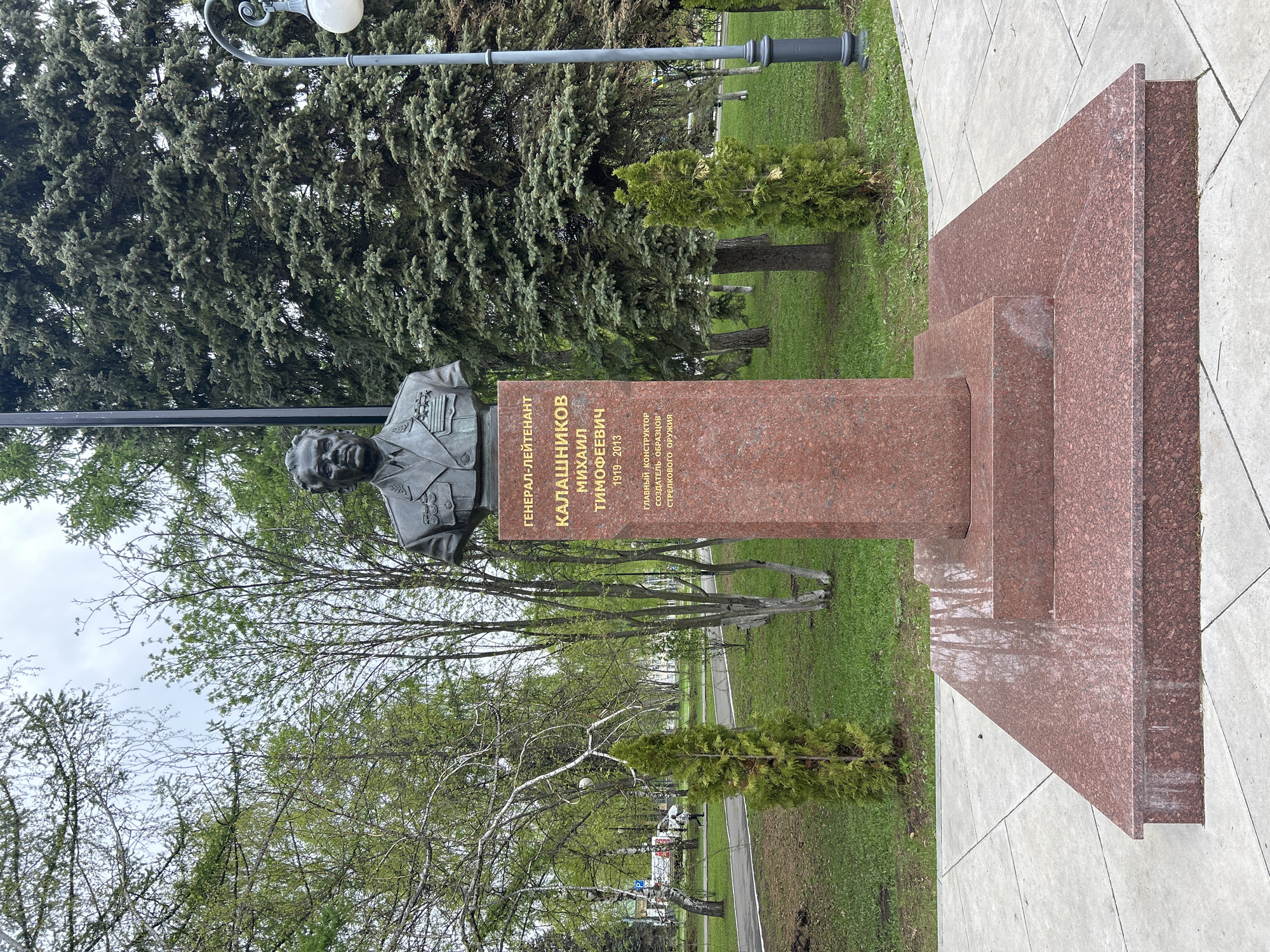 The image shows a bronze bust of Mikhail Kalashnikov, a Soviet and Russian Lieutenant General and arms designer best known for creating the AK-47 assault rifle. The bust is mounted on a reddish-brown granite pedestal with Cyrillic inscriptions. The statue is located in a park in Izhevsk, Russia. The park has paved walkways and trees in the background. The day appears overcast.