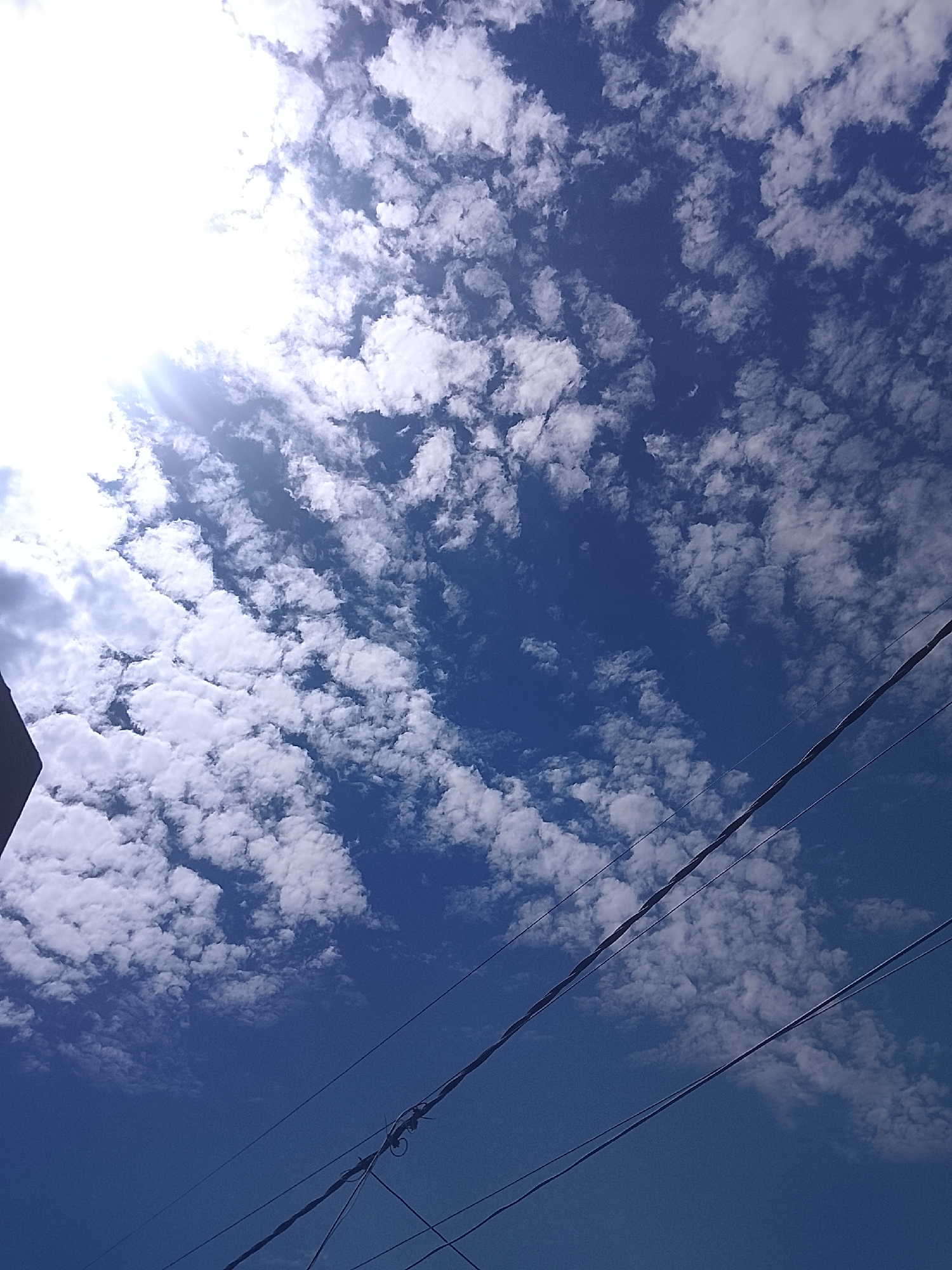 The image depicts a bright daytime sky filled with altocumulus clouds, with some areas of clear blue. The sun is visible in the upper left corner, partially obscured by clouds. Electrical wires are visible in the lower right portion of the frame. A portion of a building or structure is visible in the lower left corner. It can be assumed that this photograph was taken in Altagracia, Venezuela.