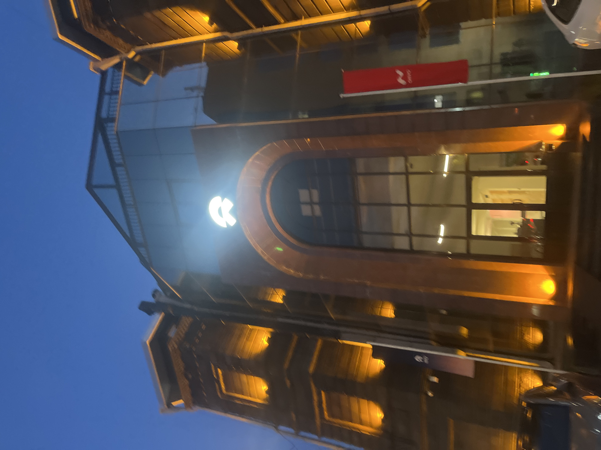 The image shows a building at night, possibly in Yerevan, Armenia, based on the logo on the building. The building has a glass facade with a large, arched entrance. There is a red sign with a white logo on the upper part of the building. Lights illuminate the exterior, and the sky is a deep blue. Cars are parked nearby.
