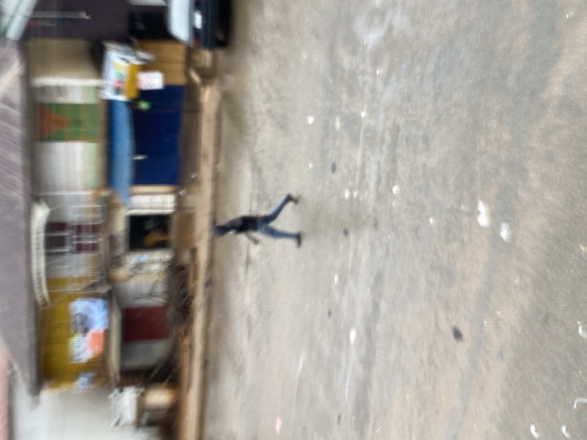A blurry overhead shot depicts a street in Bukuru, Nigeria. A person is walking along a concrete surface next to buildings. There is a car parked near the buildings. The buildings are painted in various colors.