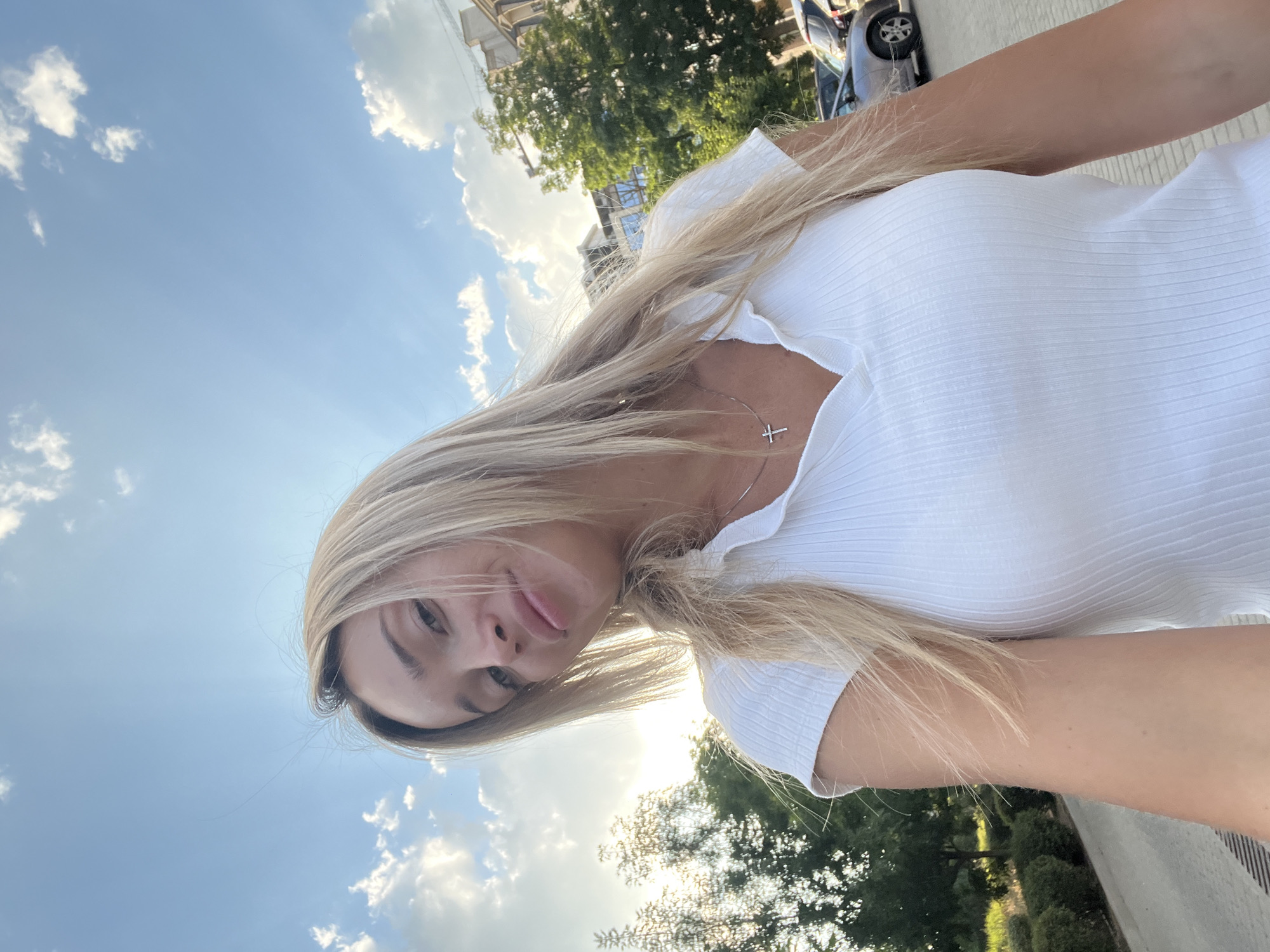 A blonde woman is taking a selfie outdoors. She is wearing a white ribbed V-neck t-shirt and a silver cross necklace. Her hair is long and blonde, and she is looking at the camera with a slight smile. The background shows a bright blue sky with white clouds, some trees, buildings, and parked cars, suggesting an urban setting. The lighting indicates it is daytime, possibly late afternoon due to the angle of the sun.