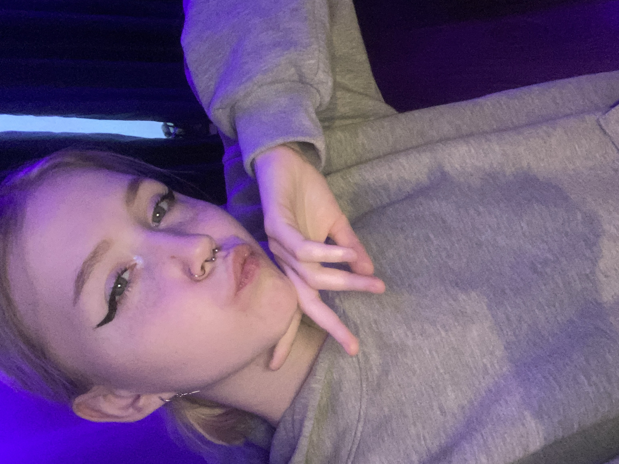 A young woman is posing for a selfie. She is wearing a gray hoodie and has visible eyeliner, a nose ring, and earrings. Her hand is positioned near her face. The background is dark, with some blue and purple lighting effects. There is no evidence to suggest this image was taken in Izhevsk, Russia.