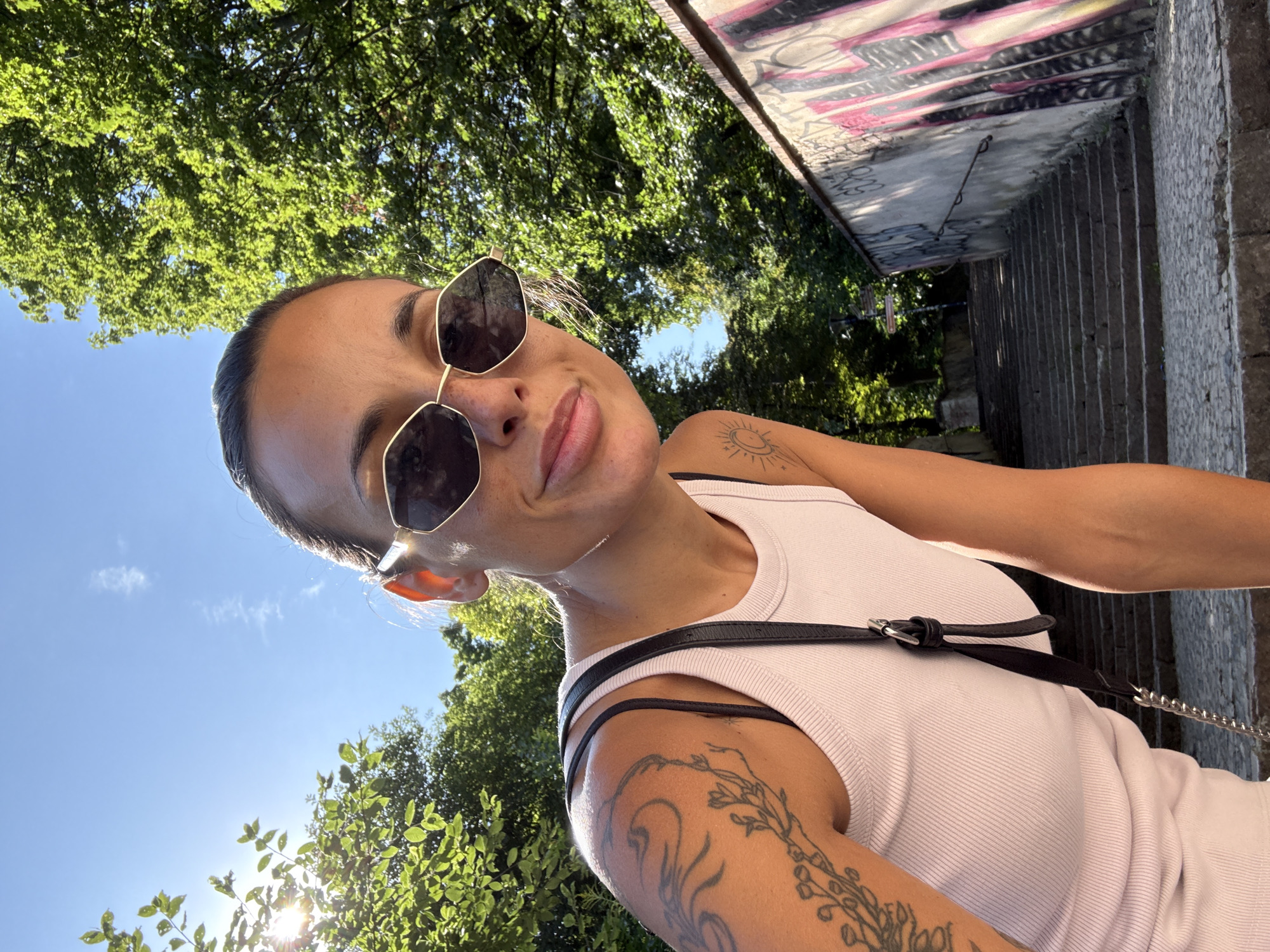 A woman is taking a selfie outdoors on a sunny day. She is wearing a light pink tank top and large, hexagonal sunglasses. She has tattoos on her left arm and shoulder, including a floral design and a sun. A black strap with a silver chain is visible across her chest, part of a bag. She is looking at the camera with a slight smile. Behind her, there are stairs leading upwards, flanked by a stone wall covered in graffiti on the right. Lush green trees and a bright blue sky are visible in the background, with sunlight streaming through the leaves. The location is a park in Prague, Czechia.
