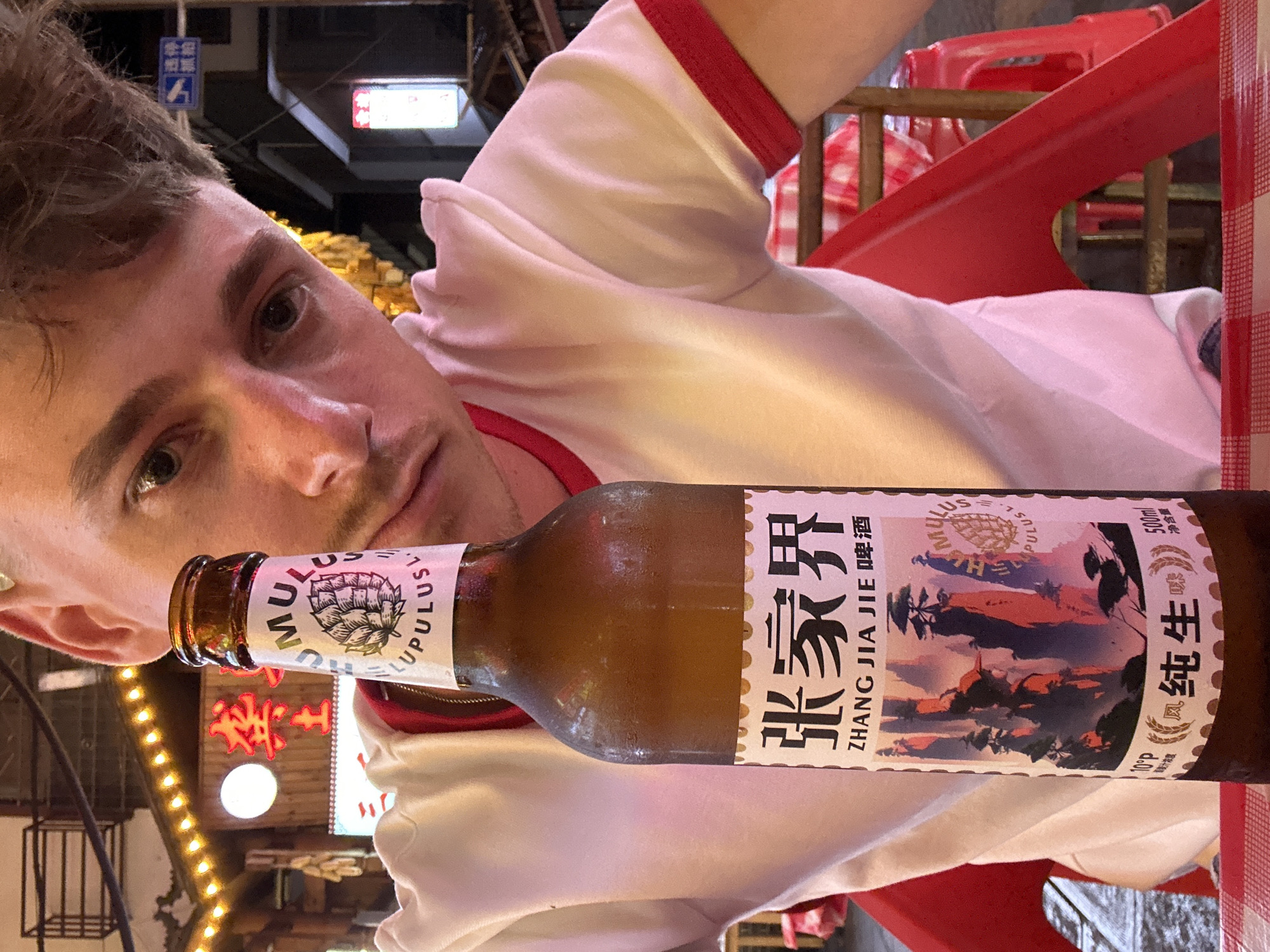 A light-skinned man, angled from the left, holds a bottle of Zhang Jia Jie beer, likely outdoors in Zhangjiajie, China, as indicated by the beer brand and Chinese signage in the background. He wears a white t-shirt with red trim. The bottle label is partially obscured by condensation. A red plastic chair is visible. The background features illuminated signs and decorative lighting.