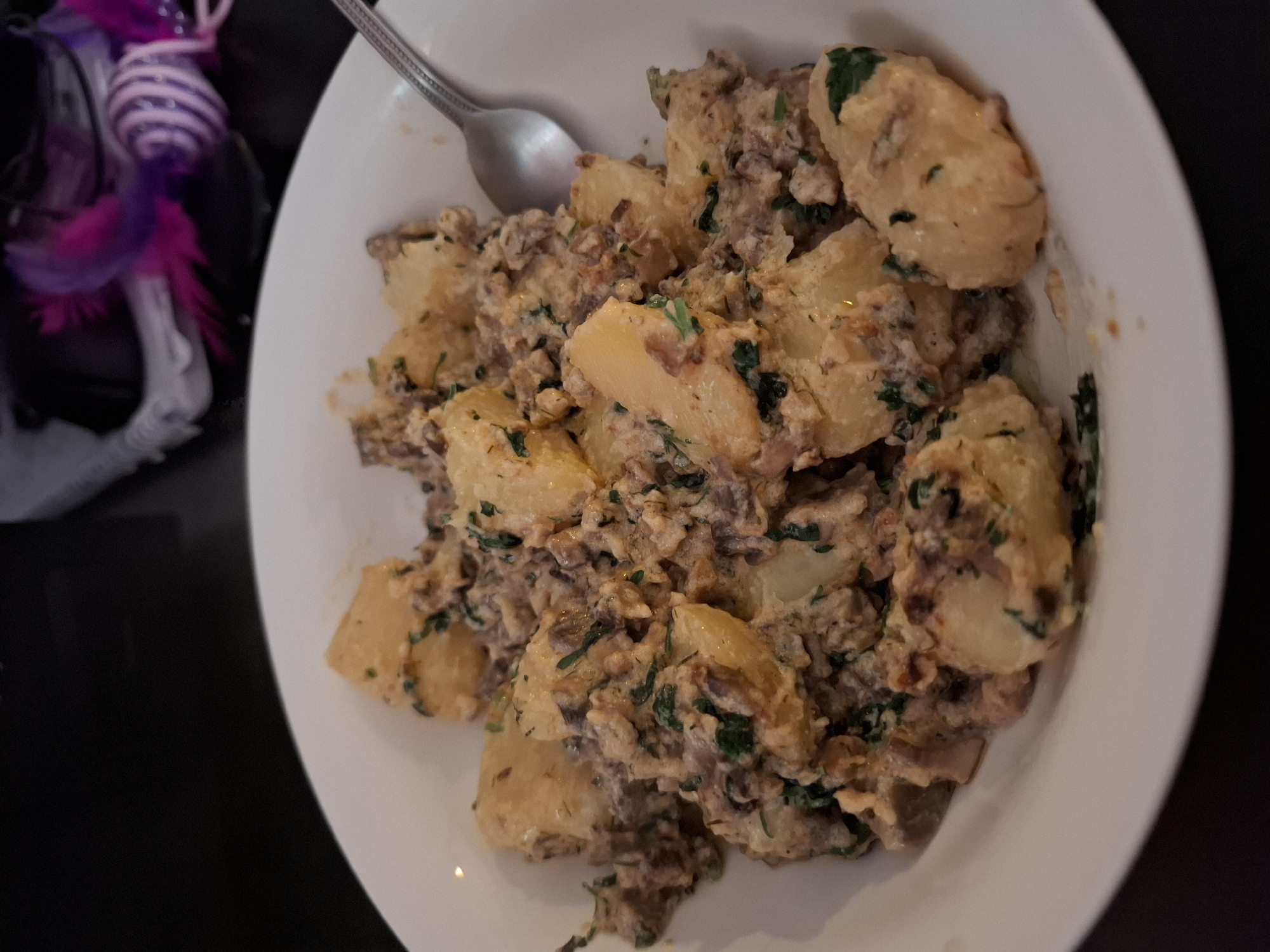 A close-up, overhead shot shows a white, round bowl filled with a potato and meat dish. The potatoes are cut into large, irregular chunks and are coated in a creamy, light brown sauce mixed with finely crumbled meat and chopped green herbs. A silver spoon rests on the right side of the bowl, partially submerged in the food. The background is dark and out of focus, with hints of purple and white objects on the right side, possibly a toy or decoration. The scene appears to be indoors, likely in a dining setting. There is no discernible indication of the location being in Zemun Urban Municipality, Serbia.
