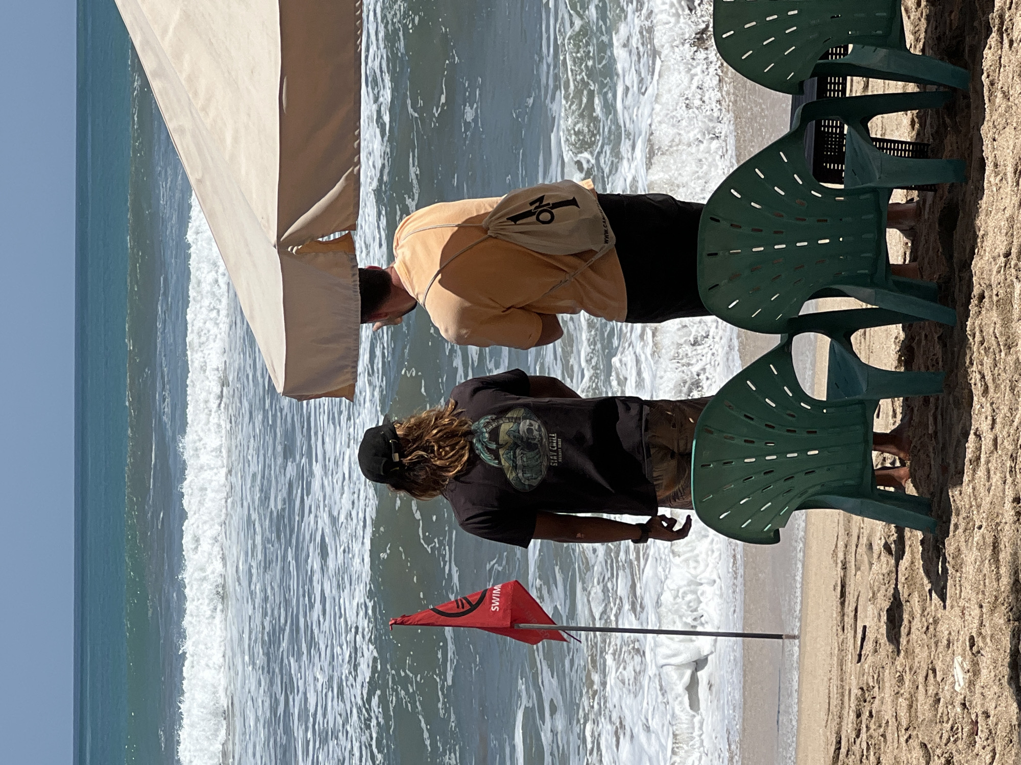 This is a vertical, eye-level, full-shot photograph of two men standing on the beach in Kuta, Indonesia, looking out at the ocean. The man on the left is wearing a black t-shirt with a graphic on the back and a black baseball cap. He has long, blonde hair. The man on the right is wearing an orange t-shirt and black shorts, with a beige drawstring bag slung over his shoulder. They are standing near a red flag with "SWIM" written on it. In the foreground, there are three green plastic beach chairs and a small black table. The ocean in the background has moderate waves, and the sky is clear and blue. The sun is shining brightly, casting shadows on the sand.