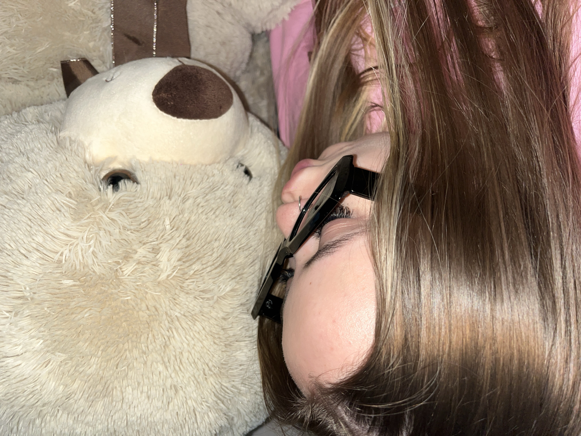 A close-up image shows a person lying on their side, possibly sleeping, next to a large, plush teddy bear. The person has brown hair with blonde highlights, is wearing black-rimmed glasses, and has a pierced nose. The teddy bear is primarily white with a brown nose and brown ears. The background appears to be soft bedding, with a hint of pink visible. The location is not discernible from the image.