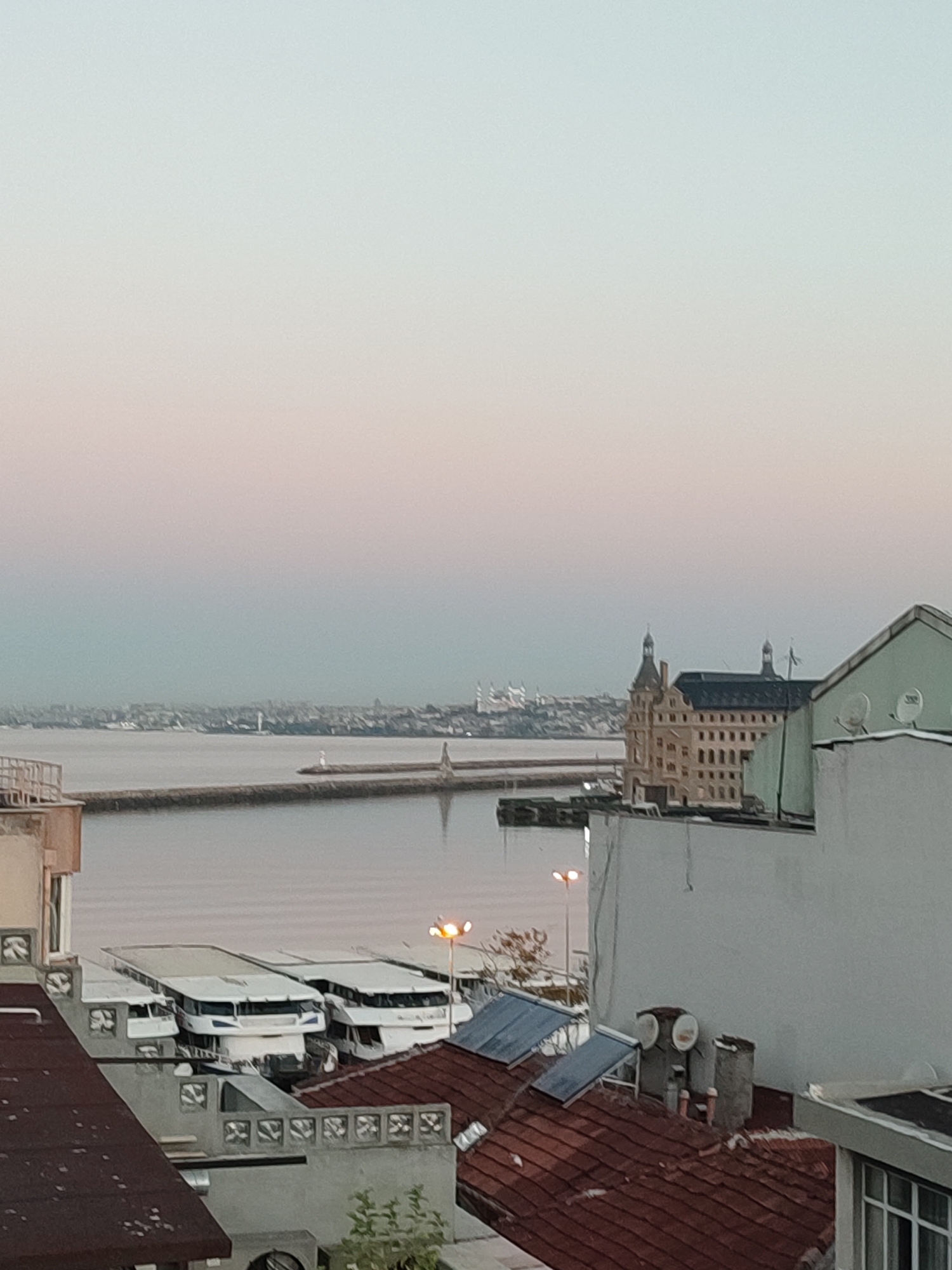 The image shows a view of the Kadıköy waterfront in Istanbul, Turkey, likely during twilight hours. In the foreground, there are rooftops of buildings, including some with red tiles and others with white or light-colored facades. Several boats or ferries are docked along the waterfront. Across the water, the distant city skyline is visible, featuring numerous buildings, including what appears to be a prominent historical structure with towers. A lighthouse is situated on a breakwater extending into the water. The sky transitions from a pale pink at the horizon to a light blue overhead. The overall scene is one of a calm, urban waterfront with architectural elements and maritime activity.