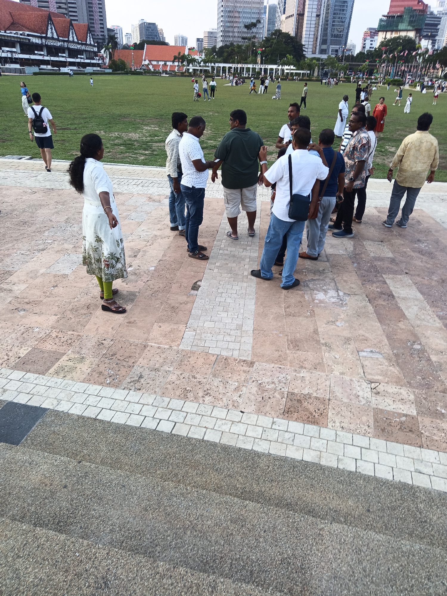 A group of people are gathered on a paved plaza in front of a large, manicured green field. In the background, tall modern buildings and some older, Tudor-style buildings are visible. The paved area is made up of large, irregular tan stones and bordered by smaller white tiles. Several individuals are standing and conversing, while others are walking or observing the surroundings. One woman in a white dress with a floral print stands facing away from the camera, while a man in a green t-shirt and shorts stands with his back to the viewer, looking out at the field. In the distance, people can be seen playing on the field. The overall setting suggests a public park or square in an urban environment. This scene is consistent with the historical Dataran Merdeka (Independence Square) in Kuala Lumpur, Malaysia, known for its blend of historical architecture and modern skyscrapers.