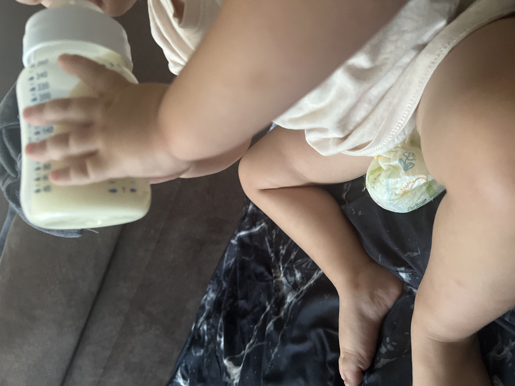 The image shows a close-up view of a baby's legs and torso, with a hand holding a baby bottle to the baby's mouth. The baby is wearing a diaper and a light-colored garment. The background appears to be a couch with a dark, marble-patterned blanket. The image does not contain any information that would allow for the determination of its location in the city of Sheptytskyi, Ukraine.