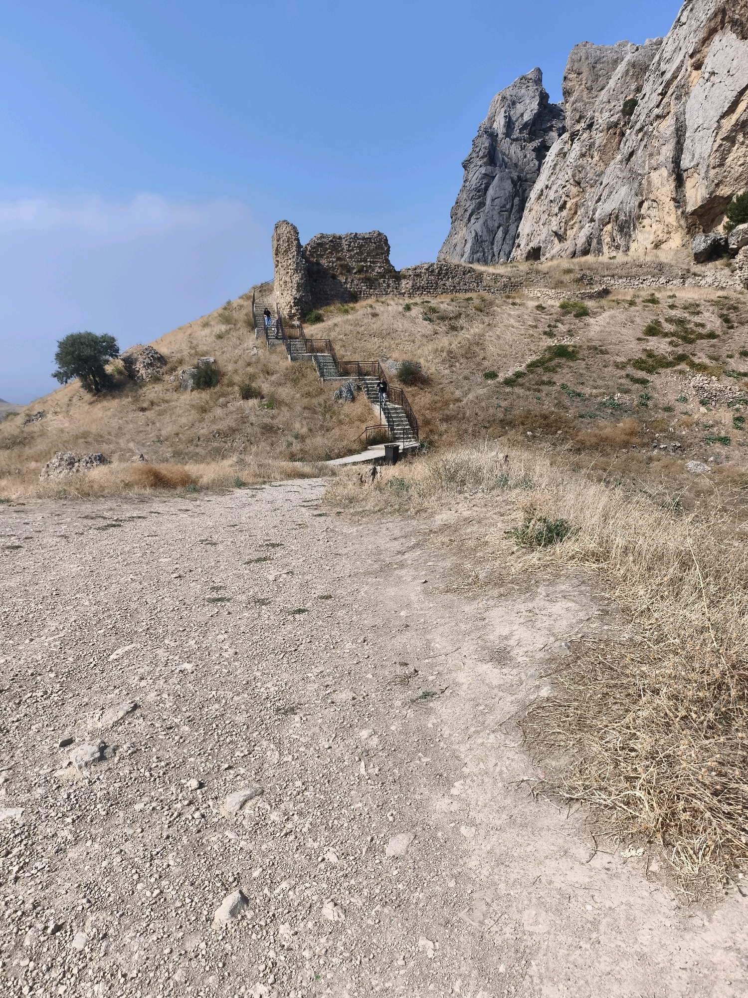 In the foreground, a path composed of gravel and dirt winds upwards. To the left and right, dry, yellowed grass covers the terrain. Further up the incline, a set of stairs with black metal railings ascends towards ancient stone ruins. A few individuals are visible on the stairs, walking. Behind the ruins, a steep, rocky mountain face dominates the background under a clear blue sky. The scene appears to be the ruins of a historical site or fortress, possibly located in Qalaşıxı, Azerbaijan, given the dry, mountainous landscape.