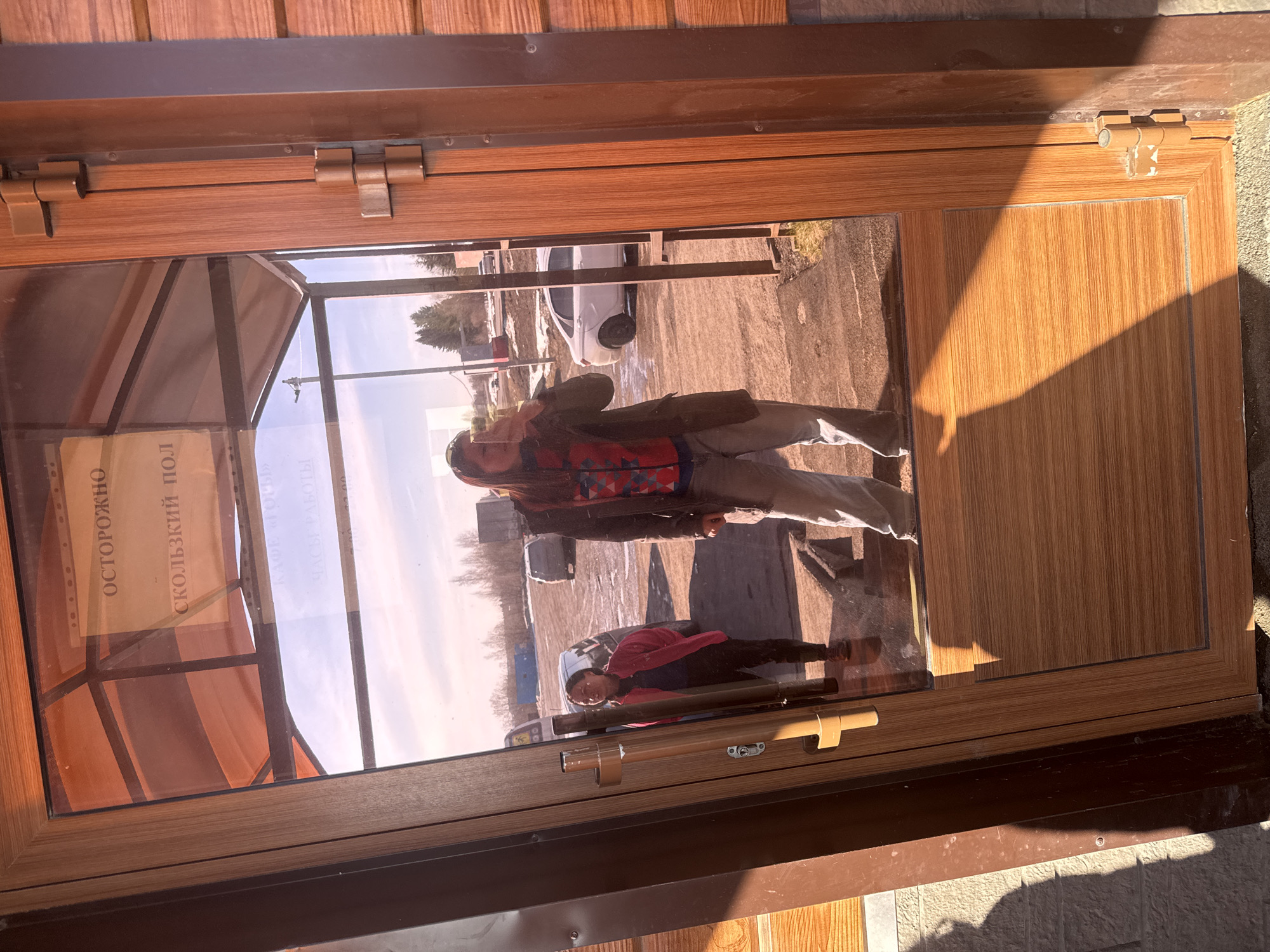 A framed window reflects a street scene in the city of Gory, Russia. Outside, two individuals stand near a car parked on a paved area. The adult is wearing a dark jacket and light-colored pants, while the child wears a pink jacket. Inside the window, a sign is visible with text in Russian, possibly warning of a slippery floor. The window frame appears to be made of wood or a wood-like material. The environment is urban with clear skies.