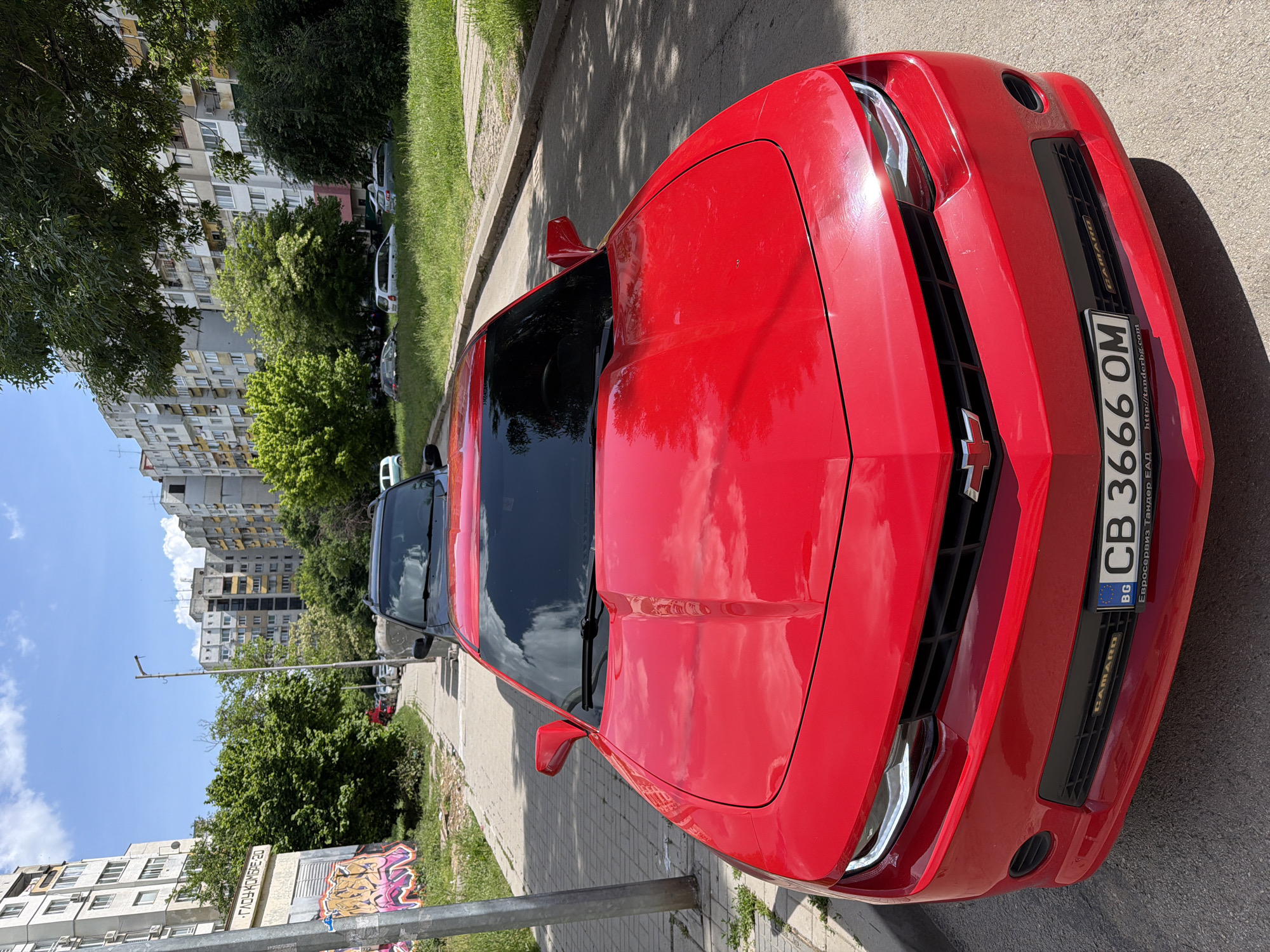 A red Chevrolet Camaro with Bulgarian license plate CB 3666 OM is parked on the side of the street in what appears to be an urban area. The street is paved, and there is a curb separating the street from a grassy area. Buildings are visible in the background, with trees lining the street. There's also a light pole with signage or graffiti on it. The day is sunny, as evidenced by the reflections on the car's hood and windshield.