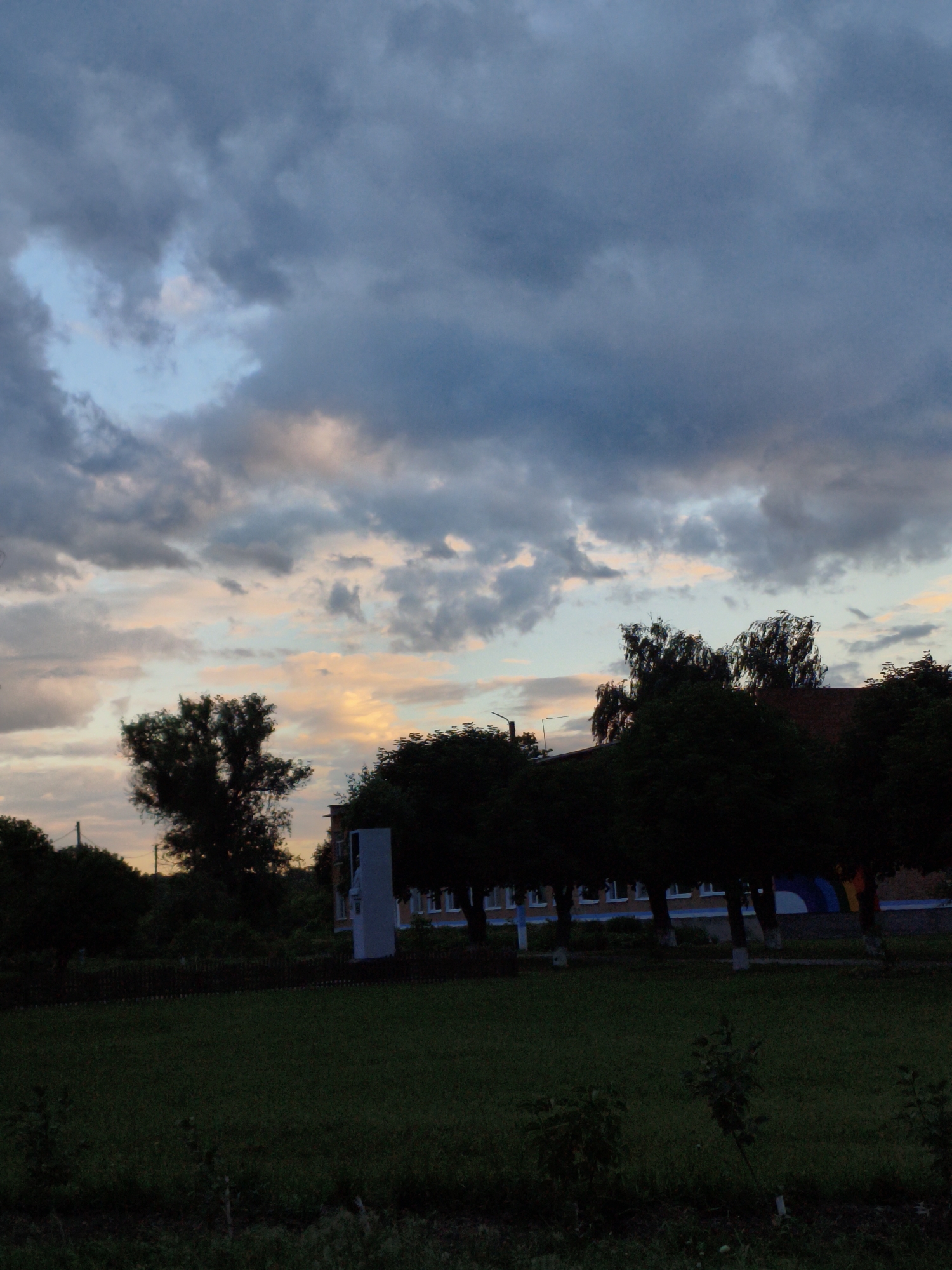 The image captures a twilight scene with a dramatic sky filled with dark, heavy clouds interspersed with patches of lighter blue and hues of pink and orange, suggesting a sunset or sunrise. In the foreground, a dark green lawn stretches out, with some small plants and shadows visible. Behind the lawn, a line of trees, silhouetted against the sky, forms a dense barrier. To the right of the trees, a building with a red-tiled roof is partially visible, and further right, a light-colored, monument-like structure stands tall. The building appears to be low-rise, with multiple windows visible on its facade, and a blue and yellow banner or mural is present on its side. The overall atmosphere is somber and slightly mysterious due to the low light and heavy cloud cover. The scene is likely located in Ustyvytsia, Ukraine, as indicated.